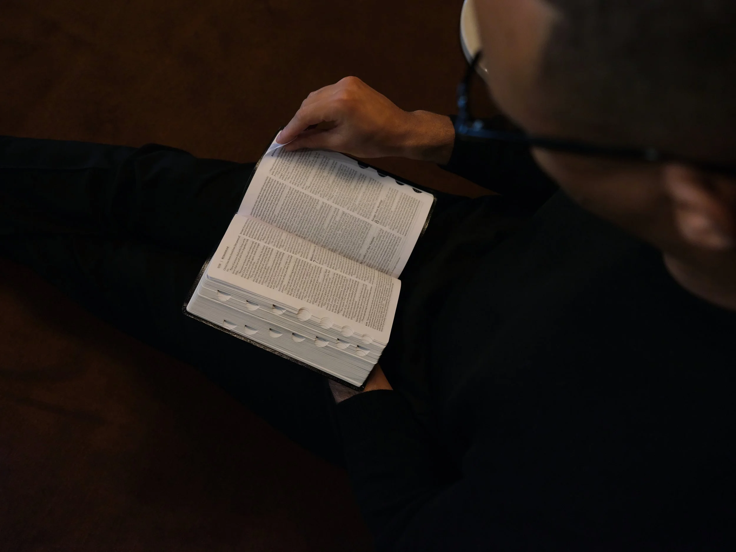 Top-down view of a person reading a Bible while sitting, with the focus on their hand holding the open Bible.