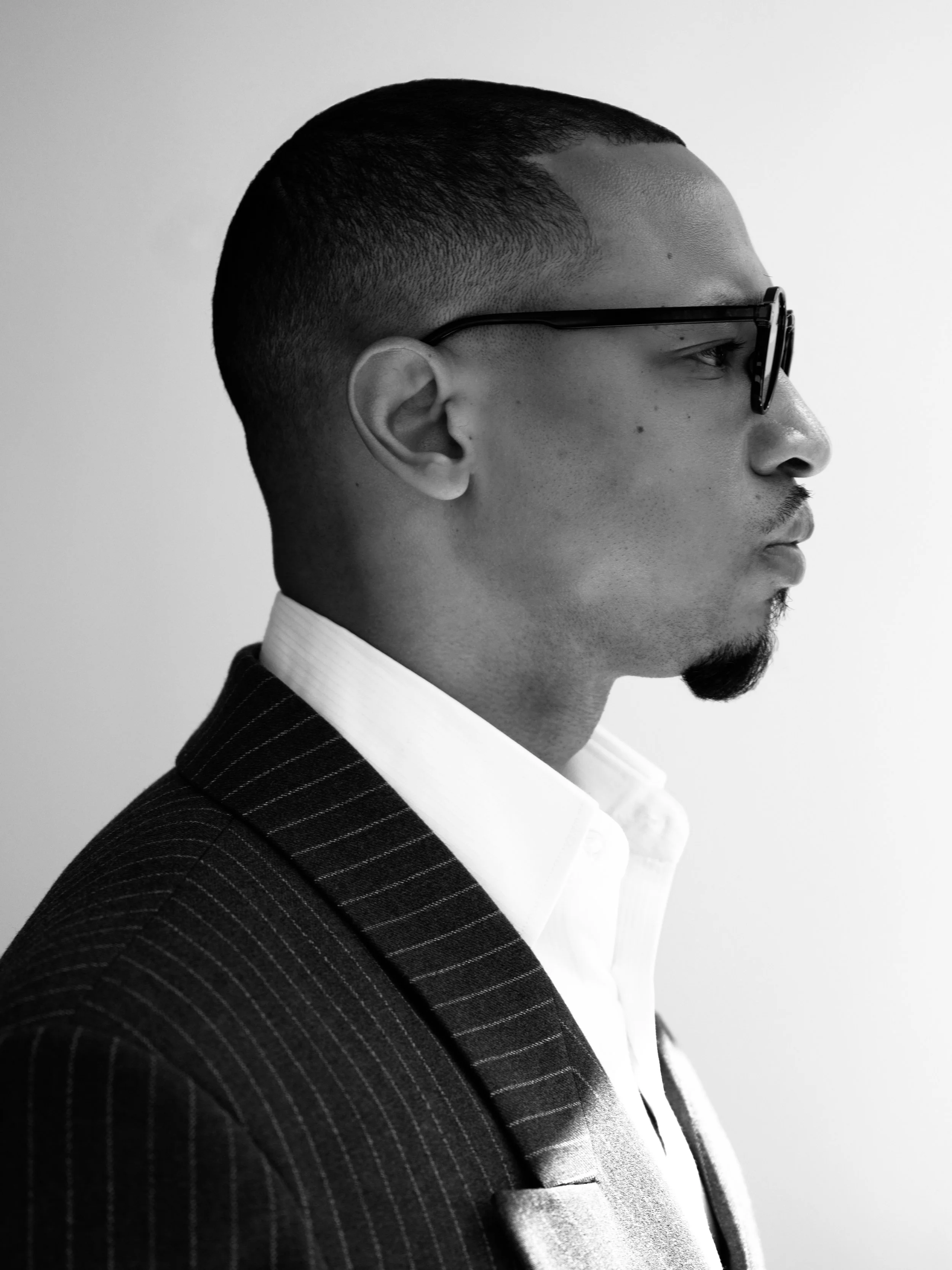 Side profile of a man in a pinstripe suit and glasses, with short hair and a goatee, against a plain background.