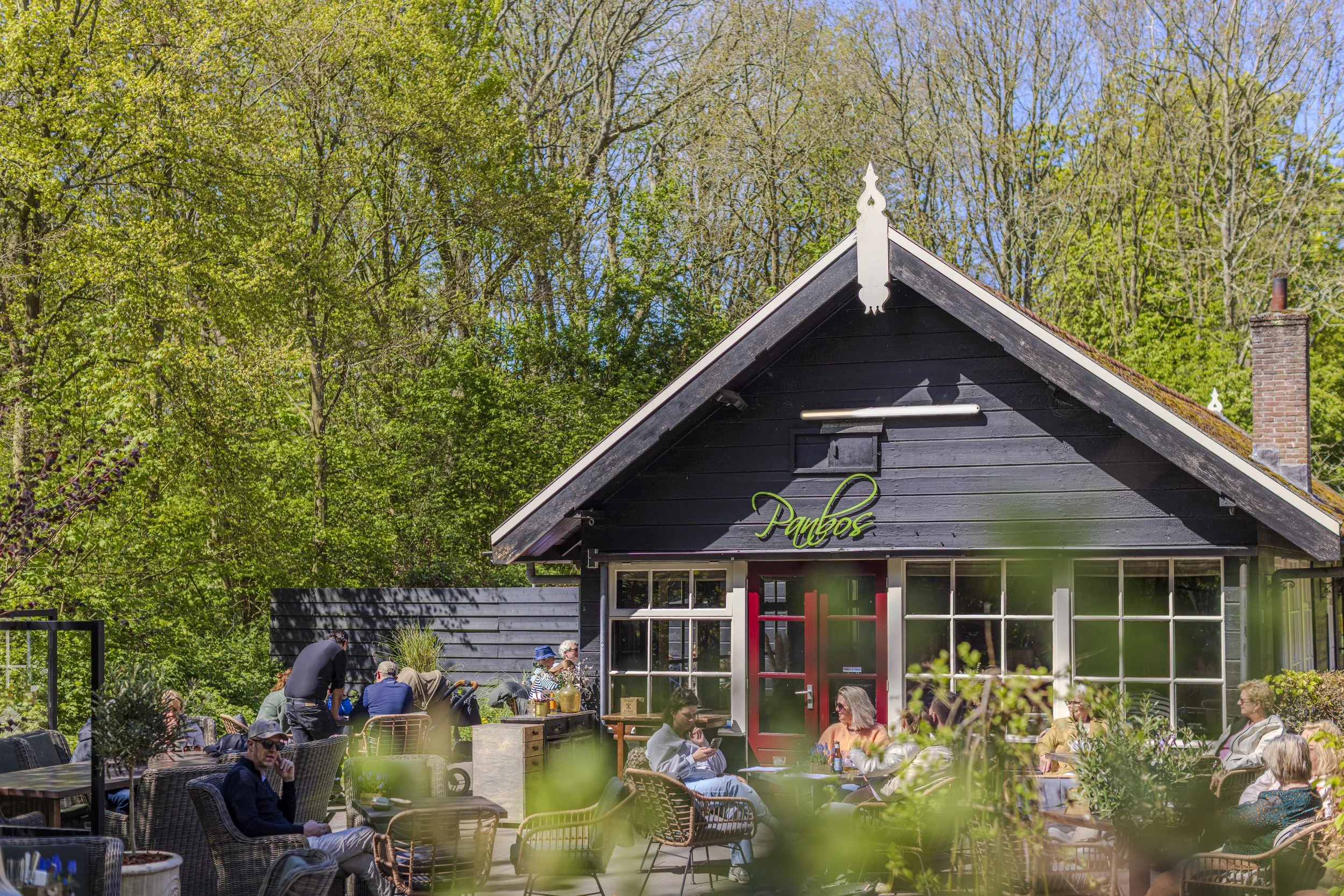 Buiten dinertafels vol mensen voor een donkerhouten gebouw met witte decoratieve elementen en grote ramen, omringd door groene bomen.