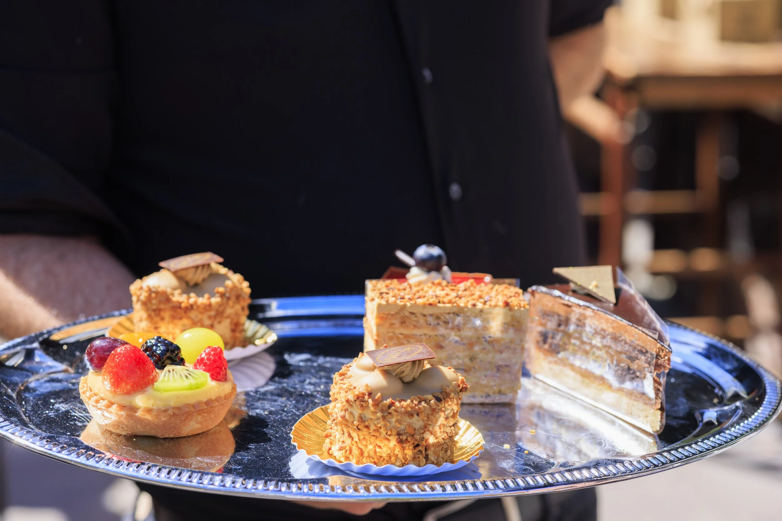 Een serveerplateau met diverse zoete desserts en fruit, gehouden door een persoon in een zwarte outfit.
