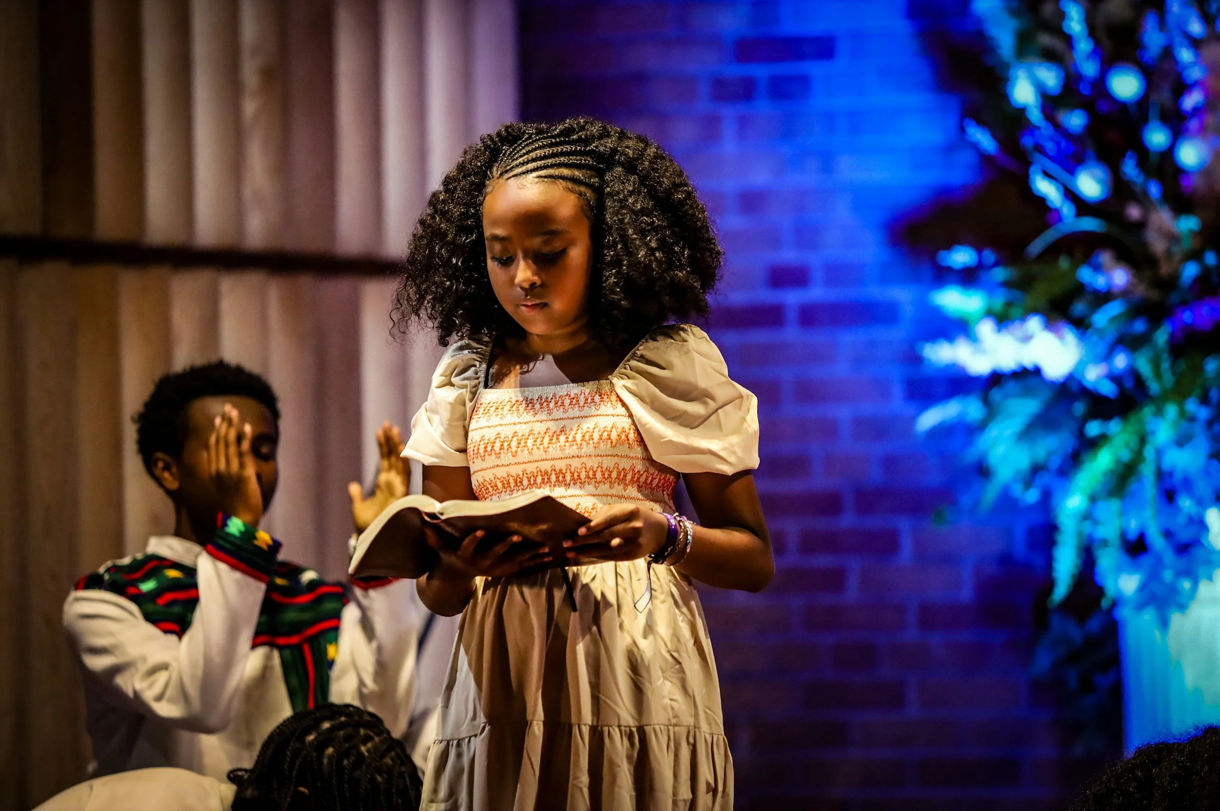 A woman with braided hair reading a book during a Christmas celebration, with a Christmas tree decorated with blue lights in the background.
