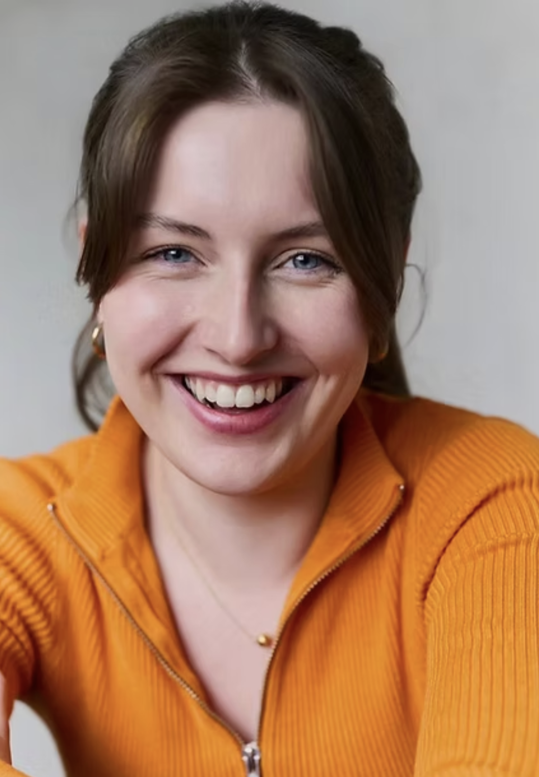 A woman with blue eyes and brown hair smiling, wearing an orange zip-up top, earrings, and a necklace.