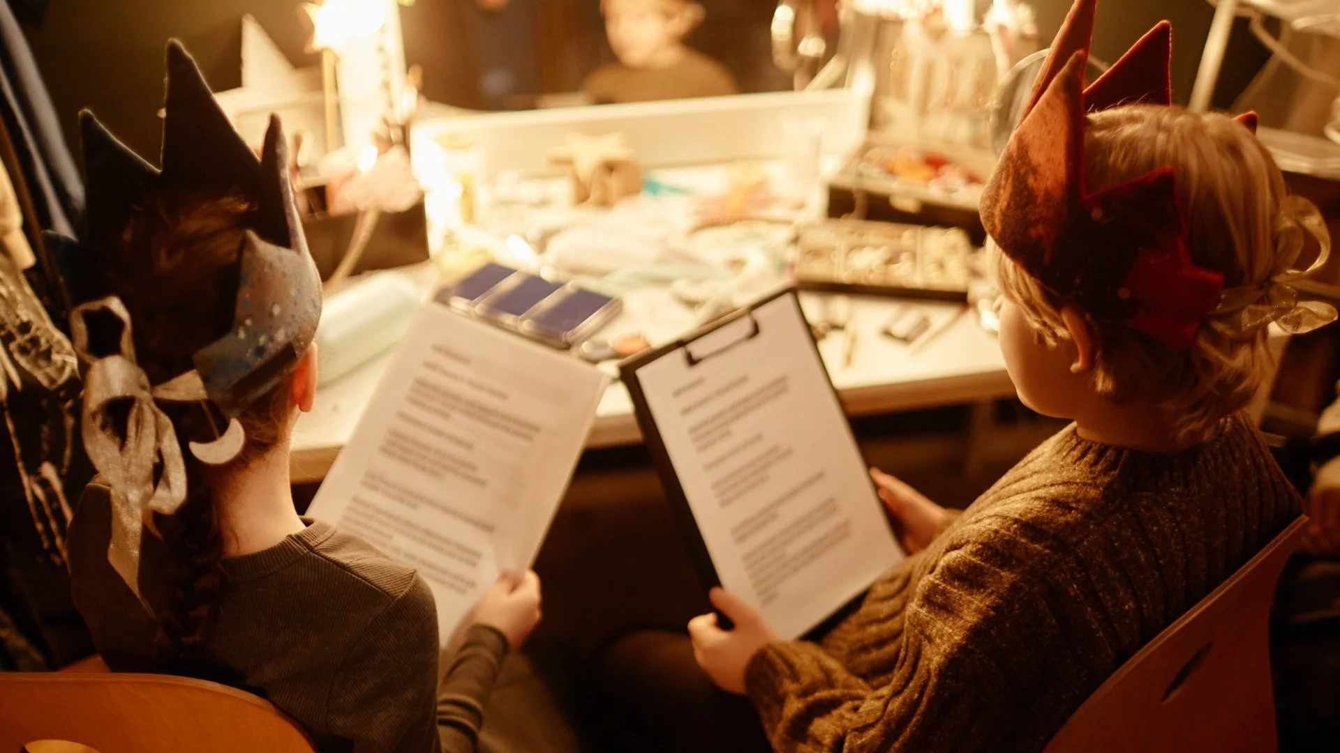 Two children sitting in theater seats, wearing Christmas-themed hats, reading menus, in a dimly lit entertainment setting.