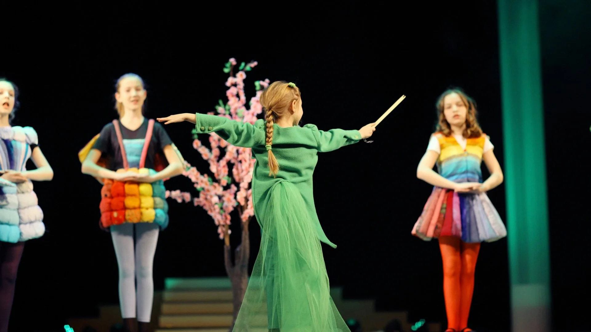 Children performing a ballet on stage, with a girl in a green costume in the center, imagining a solo.