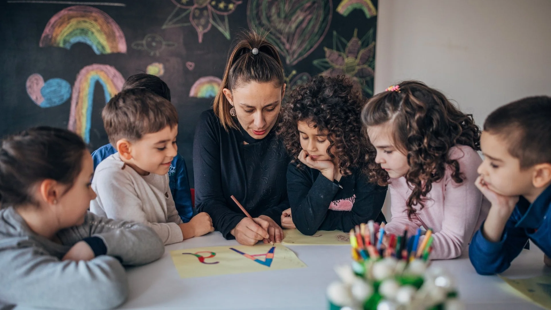 A teacher and six children gathered around a table, focusing on a drawing or writing activity; the background features a chalkboard with colorful chalk drawings of rainbows, hearts, and stars.