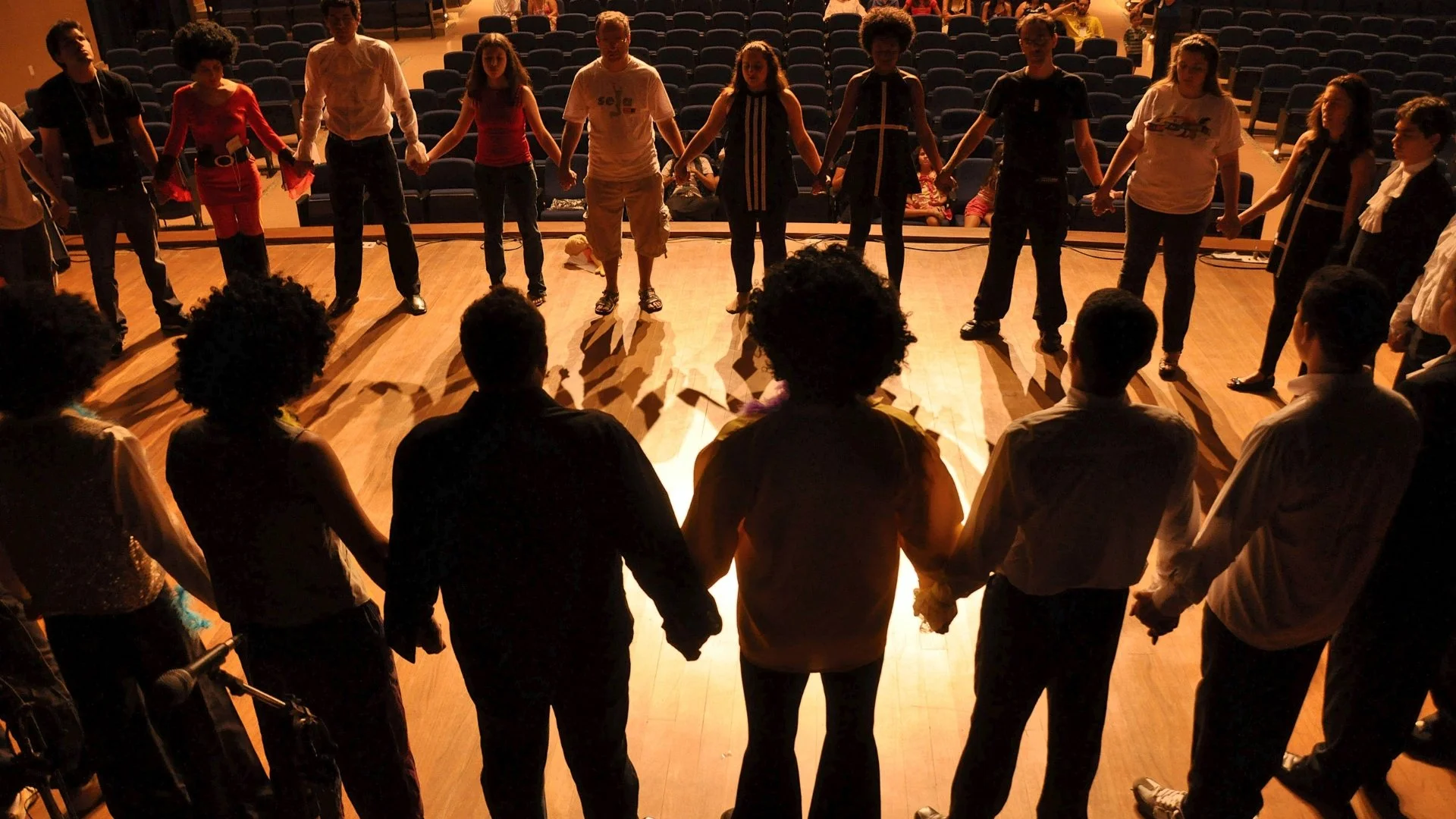 A group of people holding hands in a circle on a wooden stage, with empty chairs visible in the background.