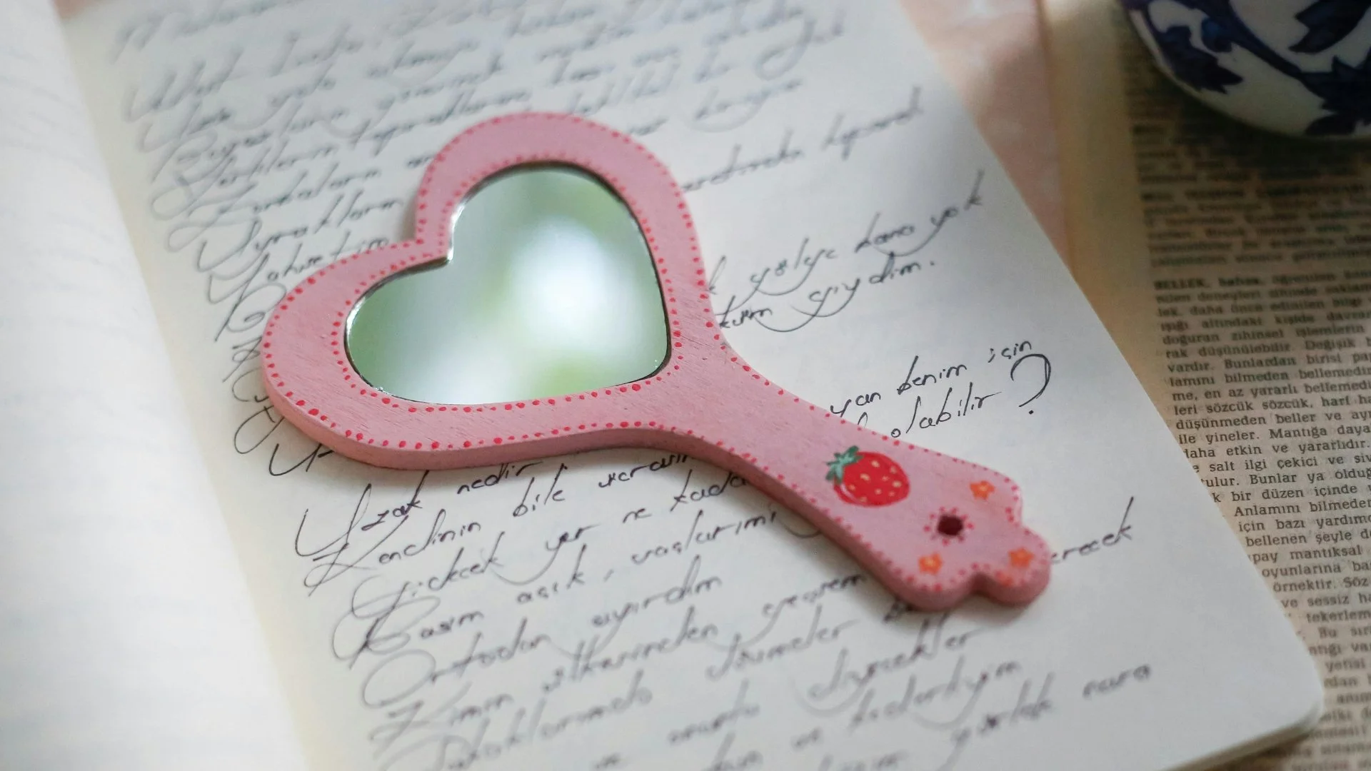 A pink heart-shaped mirror with a strawberry design on its handle, placed on handwritten notes and printed text.