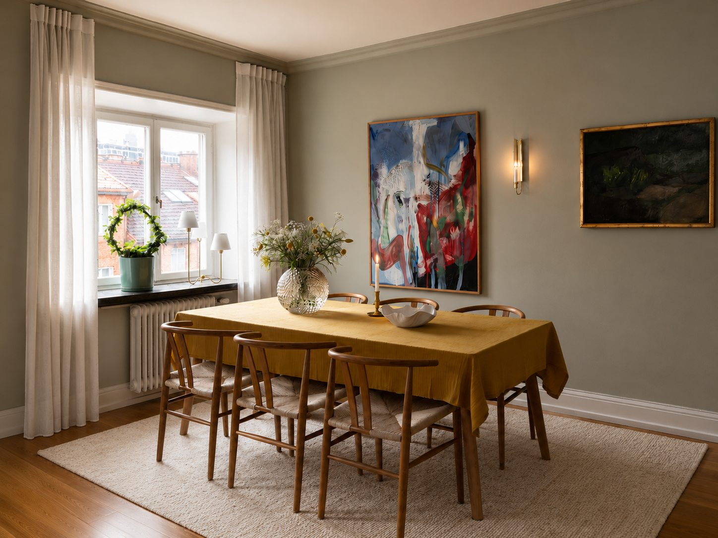 Ett matbord täckt med en gul matta, omgiven av nio trästolar med sitsar av lamull. På bordet står en stor blomsterarrangemang i ett klarglasvas, en tänd ljusstake och en skål. Ett fönster med vita gardiner och ett växt i en kruka på fönsterbrädan. Målningar på väggarna samt vägglampor. Laminatgolv och en beige matta under bordet.