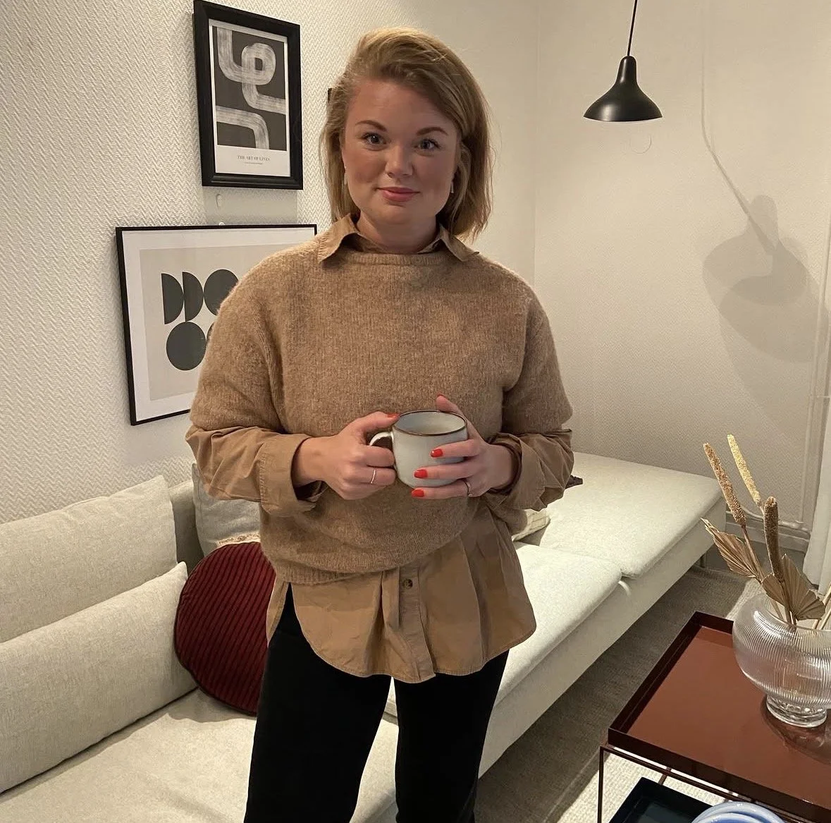 En kvinna med blont hår, iklädd beige och brunt, håller i en mugg i ett vardagsrum.