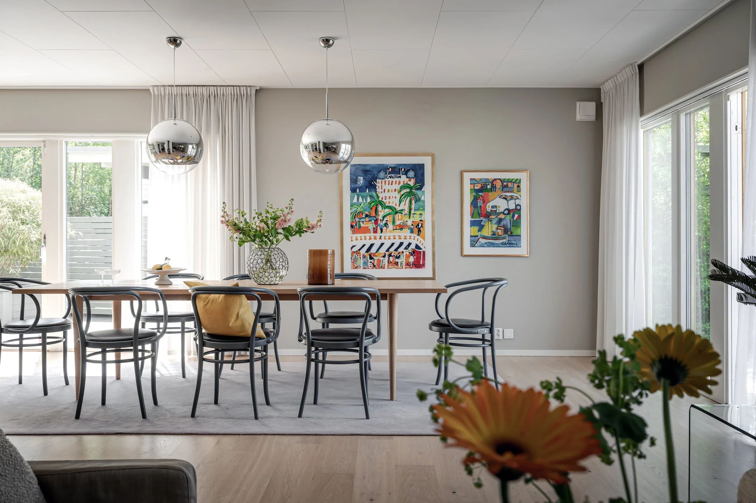 Ett modernt hem med stort matbord och stolar, konstverk på väggarna, samt stora fönster med vita gardiner som låter mycket naturligt ljus komma in.
