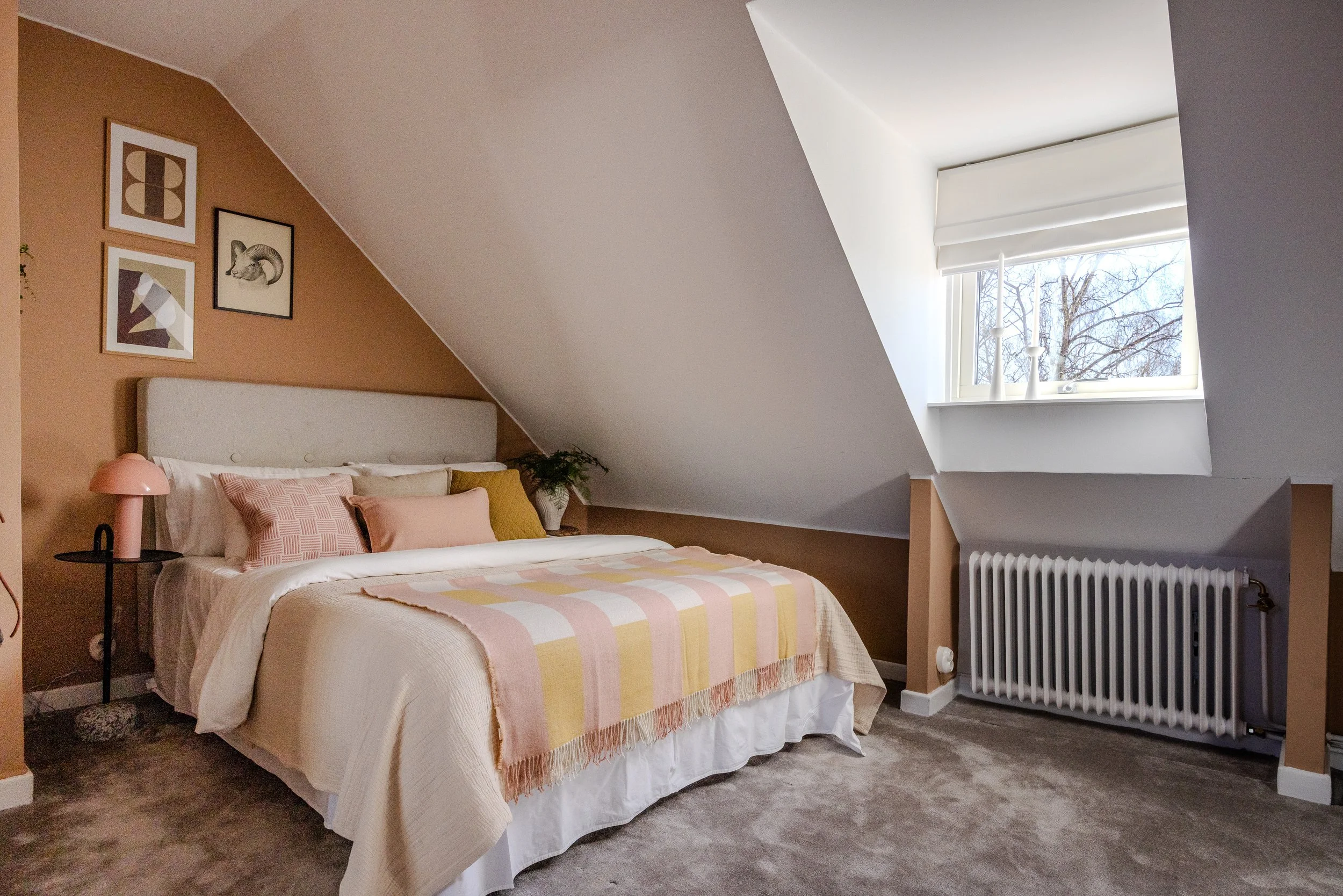 Ett sovrum med en king size-säng med ljusa sängkläder och kuddar i rosa, beige och vitt. Längs en vägg finns tre konstavelbilder och till vänster står ett litet svart sidobord med en rosa lampa. En växt i en vit kruka står vid sängens hörn. Runt sängen är det mjuk, gråbrun matta. En stor fönstervägg med vita gardiner och utsikt mot träd är på högra sidan av bilden. En vit radiator finns under fönstret.