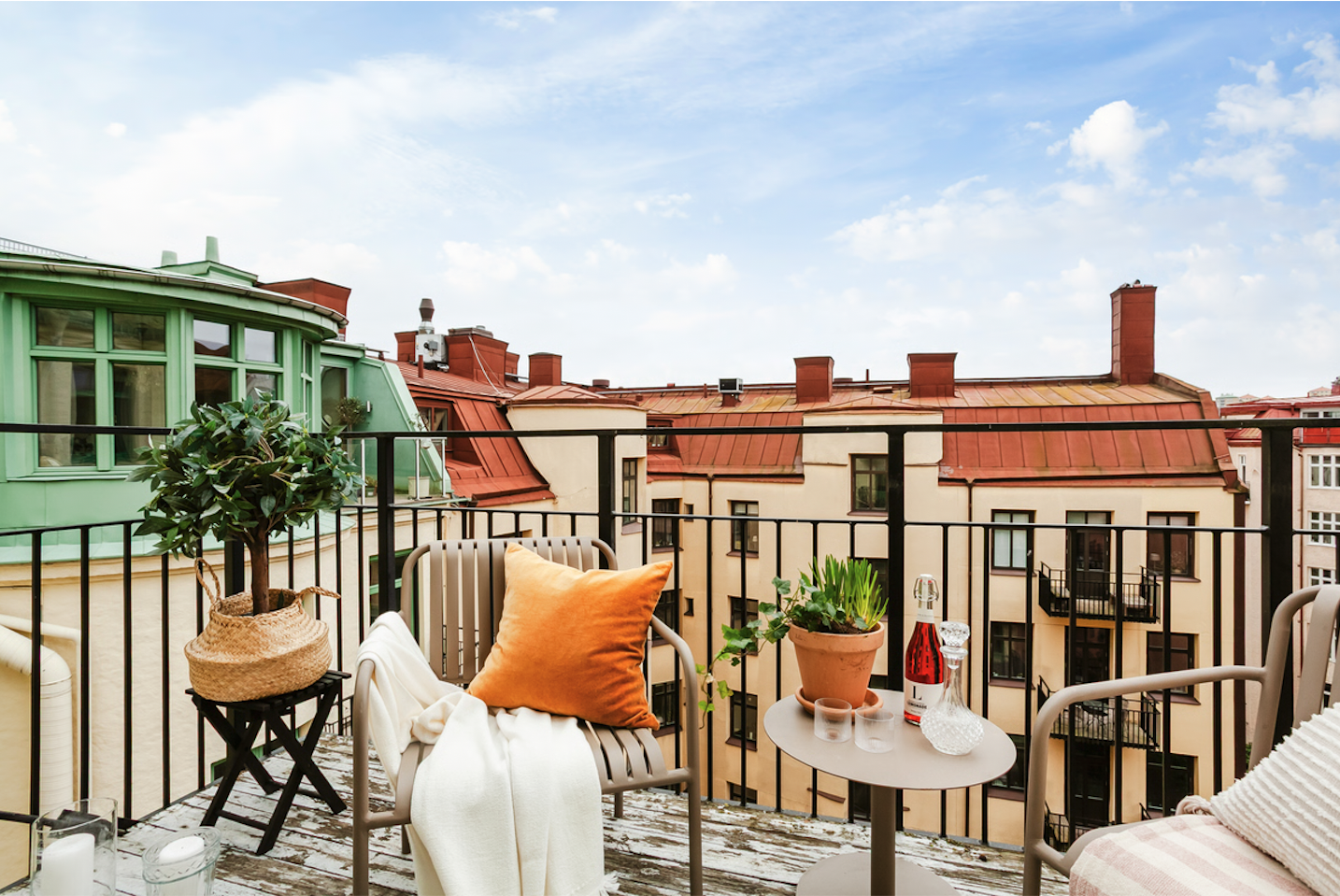 A balcony med utemöbler, en stor orange kudde, ett potted planta, en liten bord med en röd dryck, två ljuslyktor och ett glas, omgiven av byggnader med röda tak och ett blått himmel.