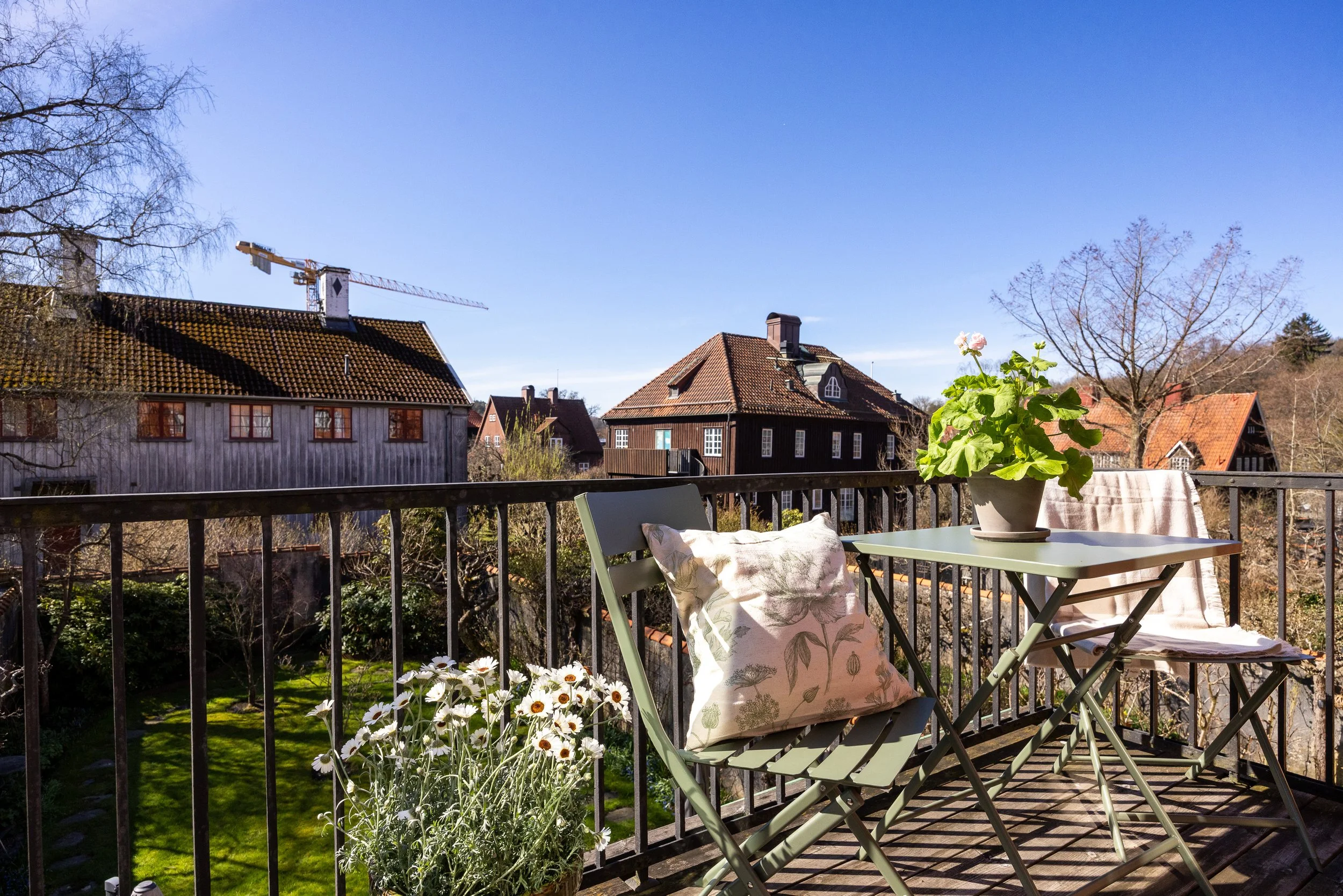 En trädgårdsvy från en balkong med ett bord, en stol med en kudde, en blomsterkruka med gröna växter och blommor, mot bakgrunden syns grannhus med tegeltak och träd mot en blå himmel.