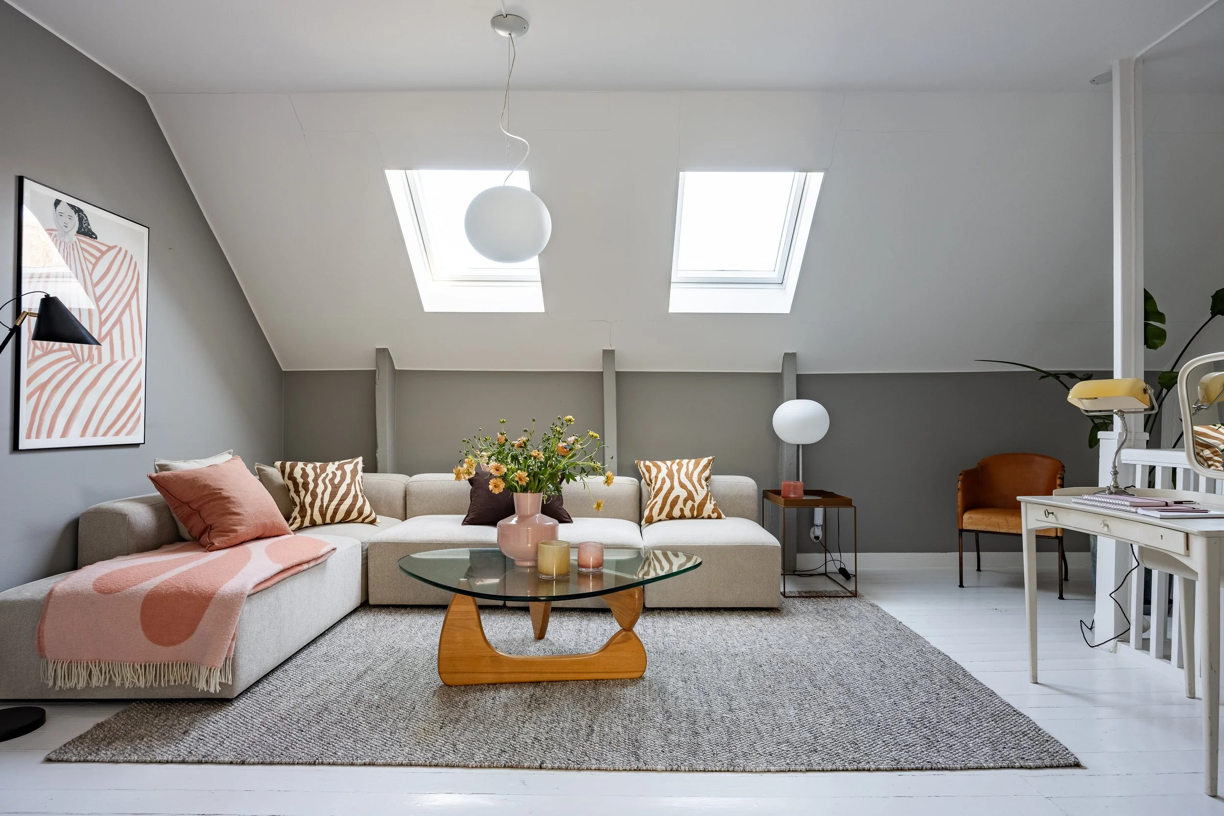 Ett vardagsrum med en ljus soffa, kuddar och en kopp med blommor på ett runt glasbord, två takfönster, väggdekoration och en golvlampa, samt ett mindre bord och stol.