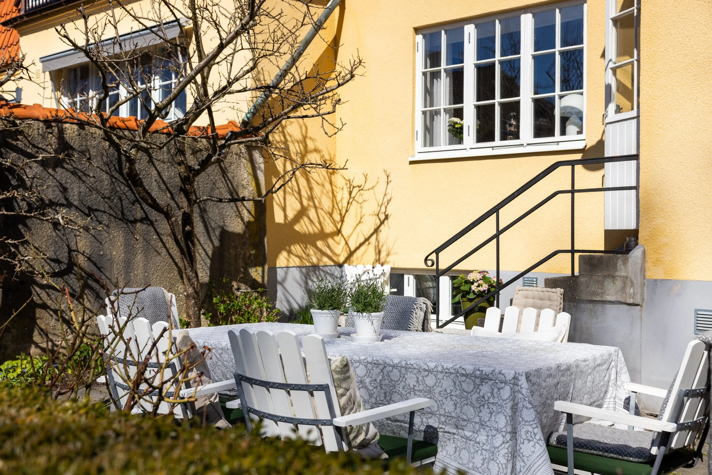 Utomhusstorbord med vita stolar, blommor i krukor, ett gult hus med stora fönster och en trappa.