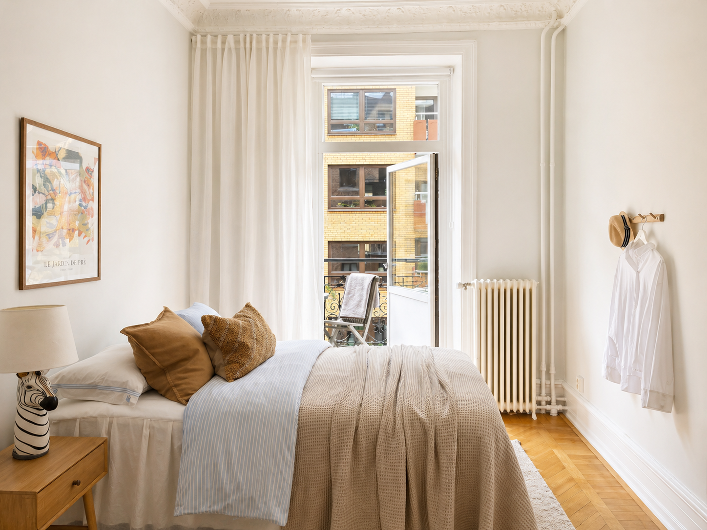 Sovrum med vita väggar, ett säng med vita och beige sängkläder, en liten nattduksbord med en zebra-formad lampa, ett fönster med ljusa gardiner som öppnar ut till en liten balkong, en stol med ett handduksdraperi, en tavla på väggen, en vit skjorta och en hatt hängande på en krok.
