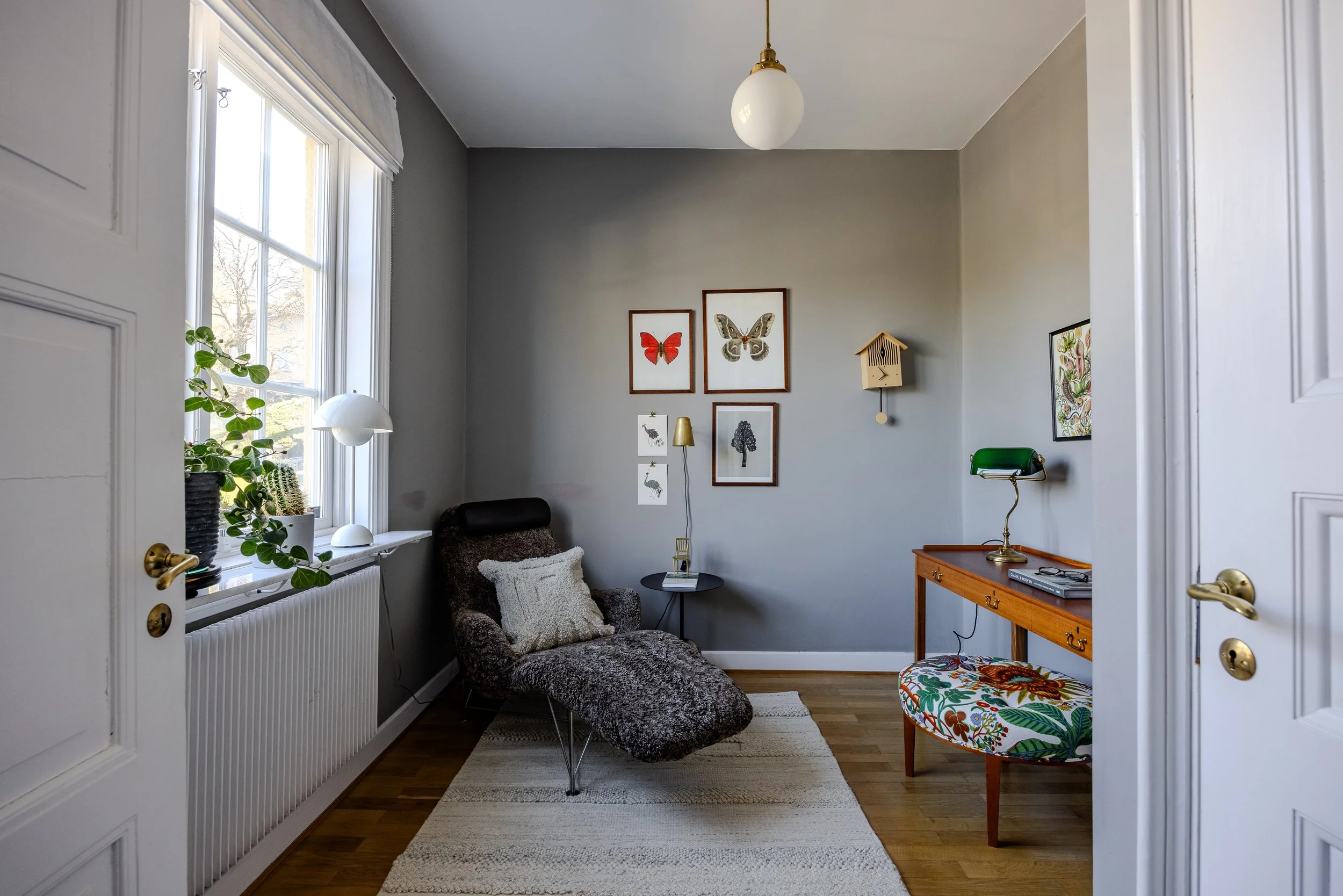 Ett mysigt vardagsrum med grå väggar. Det finns en grön vägglampa, ett svart och vitt fåtölj, en liten svart soffbord och en färgglad stol. På väggen finns fyra ramar med olika bilder av fjärilar och en väggklocka. En växt står på fönsterbrädan, som även har en vit bordslampa. Fönstret har vita gardiner.
