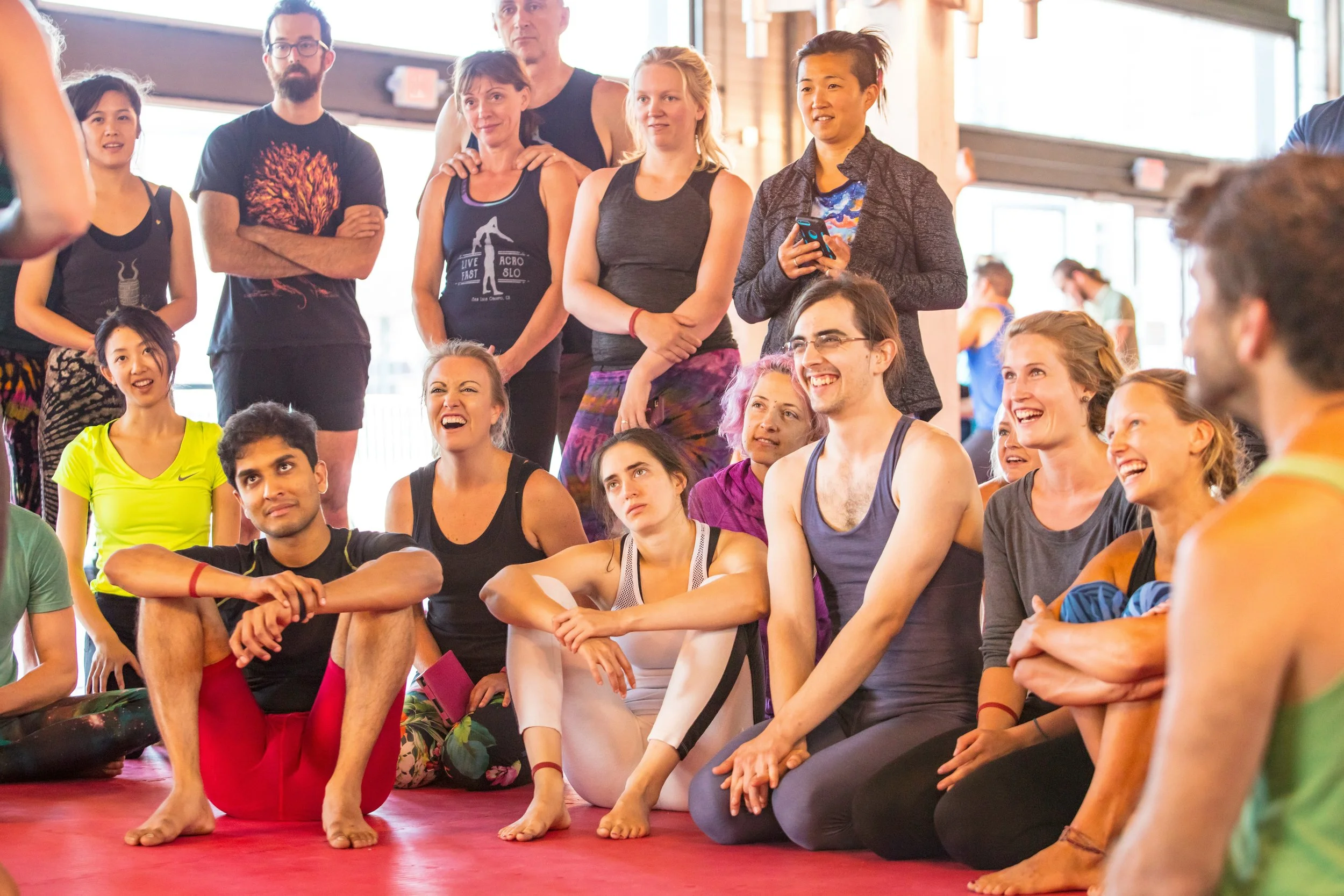 A group of people at a fitness or yoga class, sitting on a red mat, with some standing behind them, in a bright, airy studio with large windows and wooden beams.