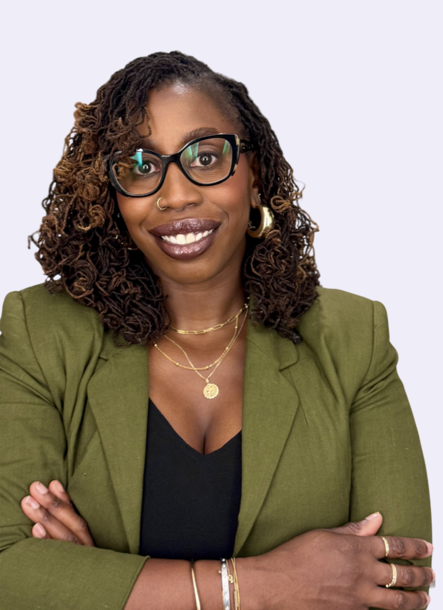 A woman with dark skin and curly brown hair wearing glasses, gold jewelry, a black top, and an olive green blazer, smiling with arms crossed.