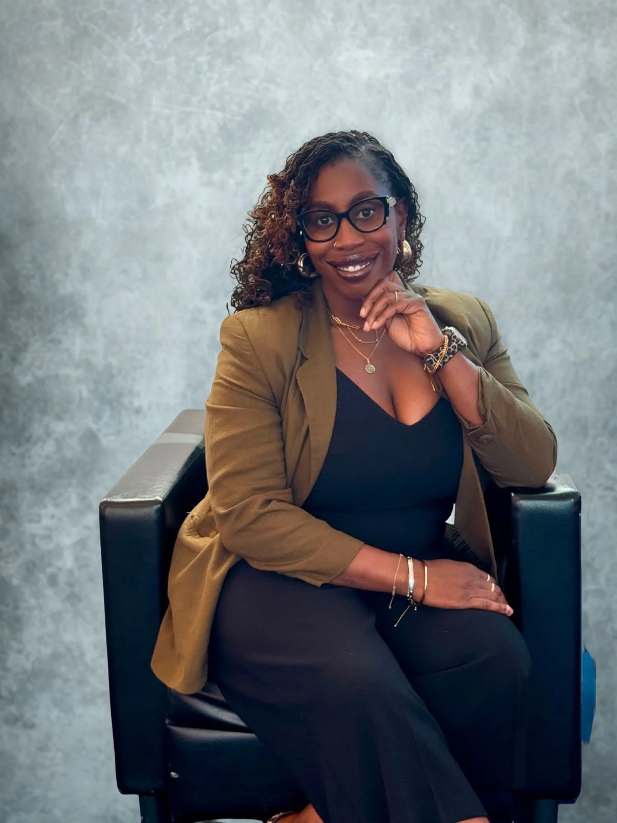 A woman with curly hair wearing glasses, a black top, and an olive green blazer sitting on a black chair against a textured gray wall.