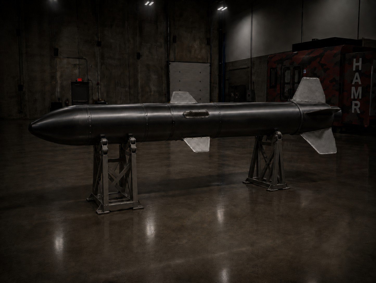 A dark-colored torpedo displayed on two metal stands inside a dimly lit exhibition room with concrete walls and wooden flooring.