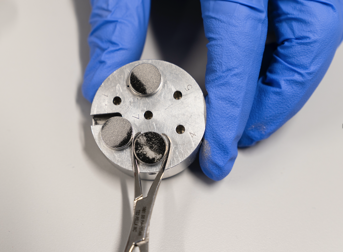 A gloved hand holding a small metal disc with three small round objects attached, one being handled with tweezers, on a white surface.