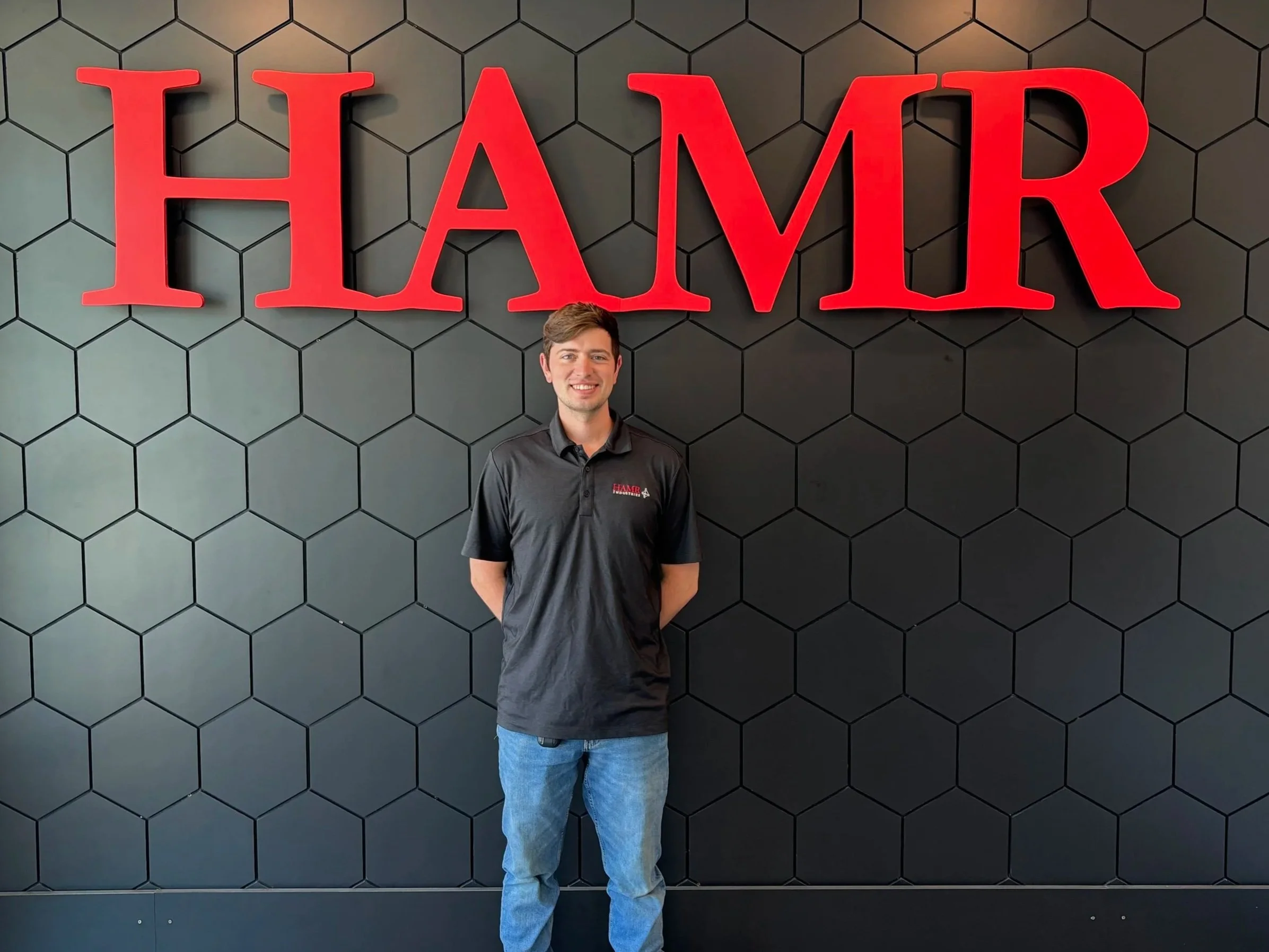 A man in a black shirt and jeans standing in front of a black wall with hexagonal tiles and a large red 'HAMR' sign