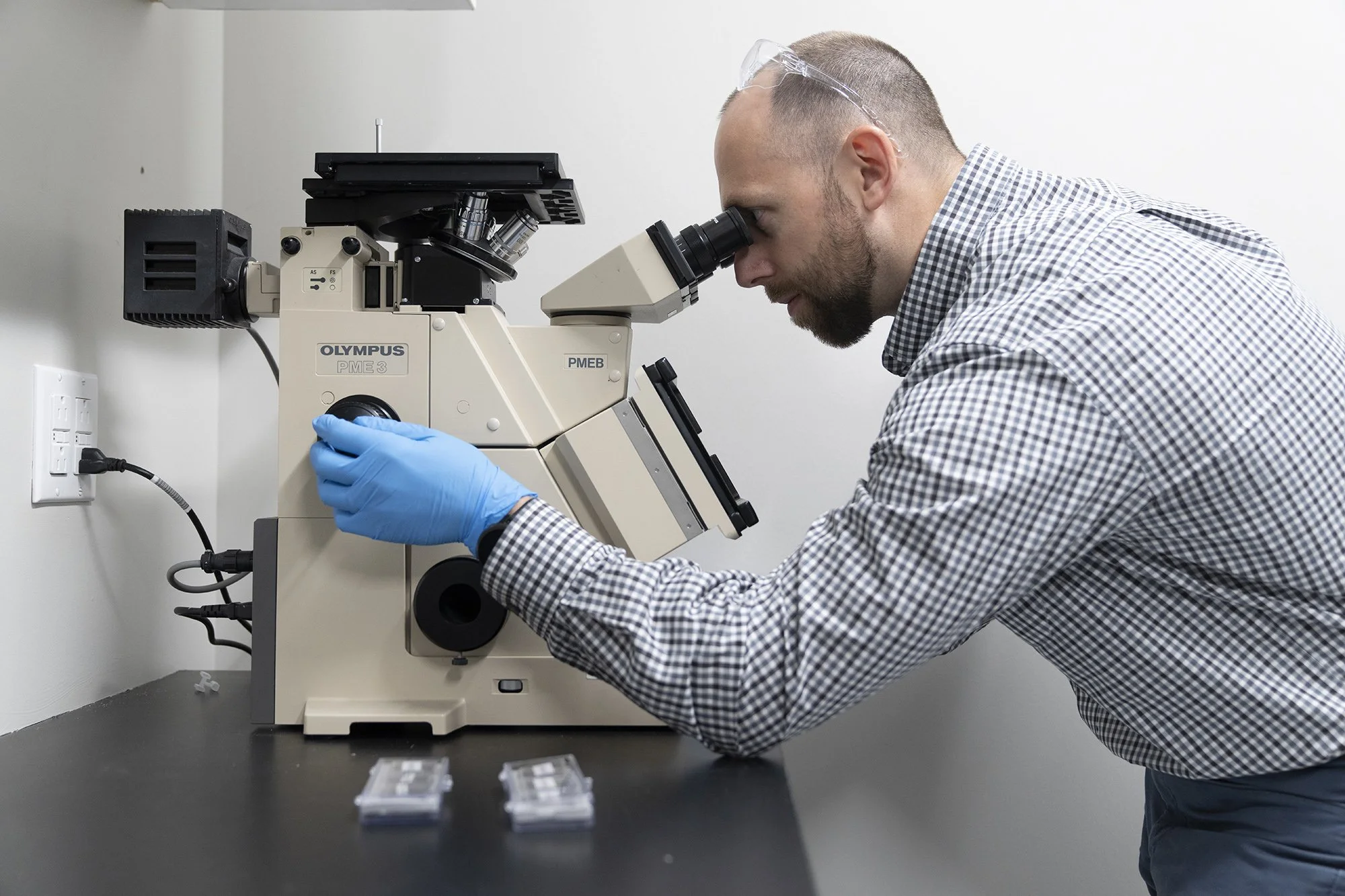 A scientist looking into a microscope in a laboratory setting, wearing blue gloves and a checkered shirt.