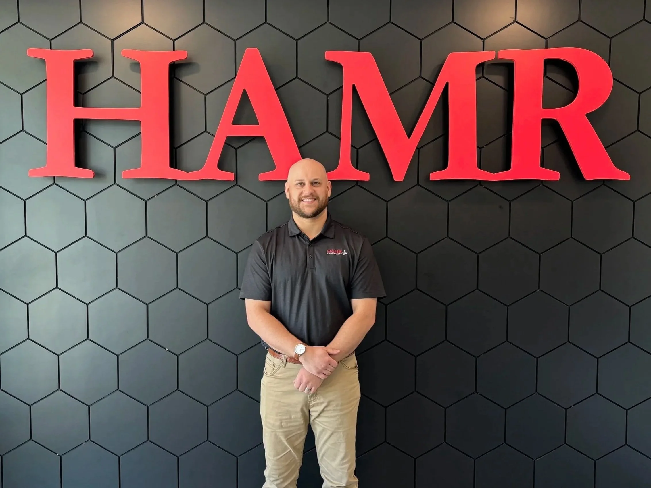 A man with a beard, wearing a black shirt and beige pants, standing in front of a black wall with a honeycomb pattern. Behind him is a large red sign that says 'HAMR'.