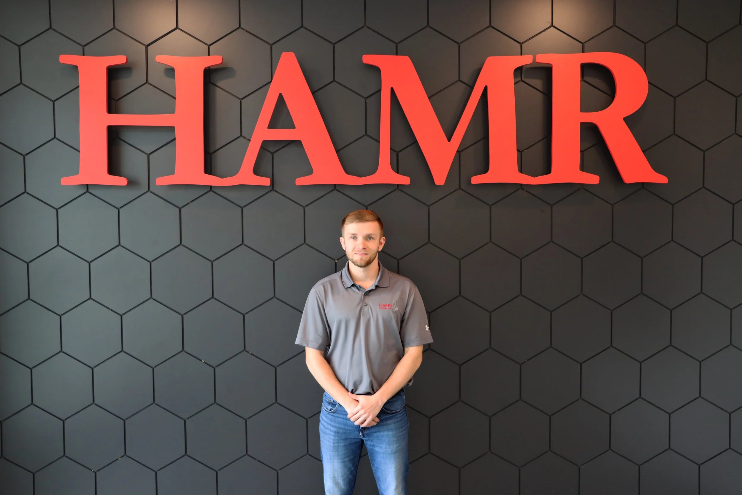 A man standing in front of a black wall with a honeycomb pattern and a large red sign that says 'HAMR'