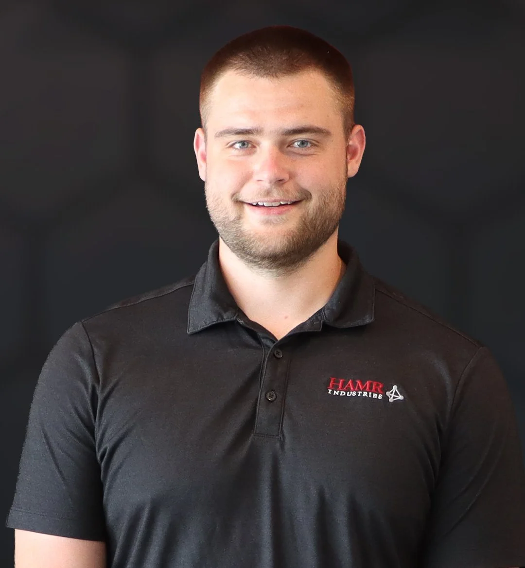 A young man with short brown hair and a beard, wearing a black polo shirt with a red and white logo that says 'HAMB INDUSTRIES' and a small white symbol, standing against a dark background.