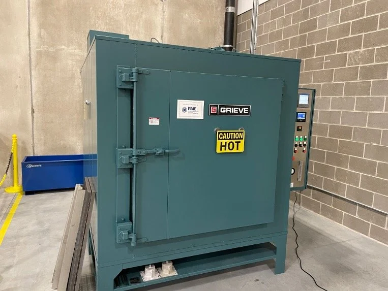 Industrial oven or heat treatment chamber with control panel, labeled with Grieve brand and caution hot sign, placed in an industrial setting with concrete floor and brick wall.