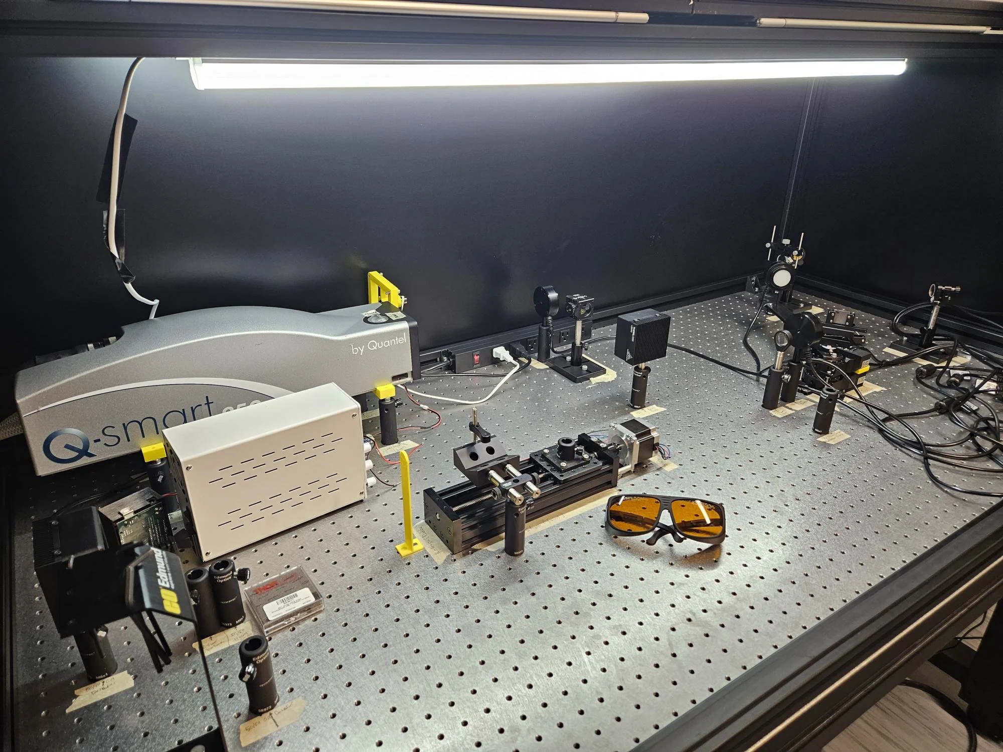 A laboratory workspace with various scientific instruments and equipment, including microscopes, optical mounts, and electronic devices, arranged on a metal perforated workbench under bright overhead lighting.