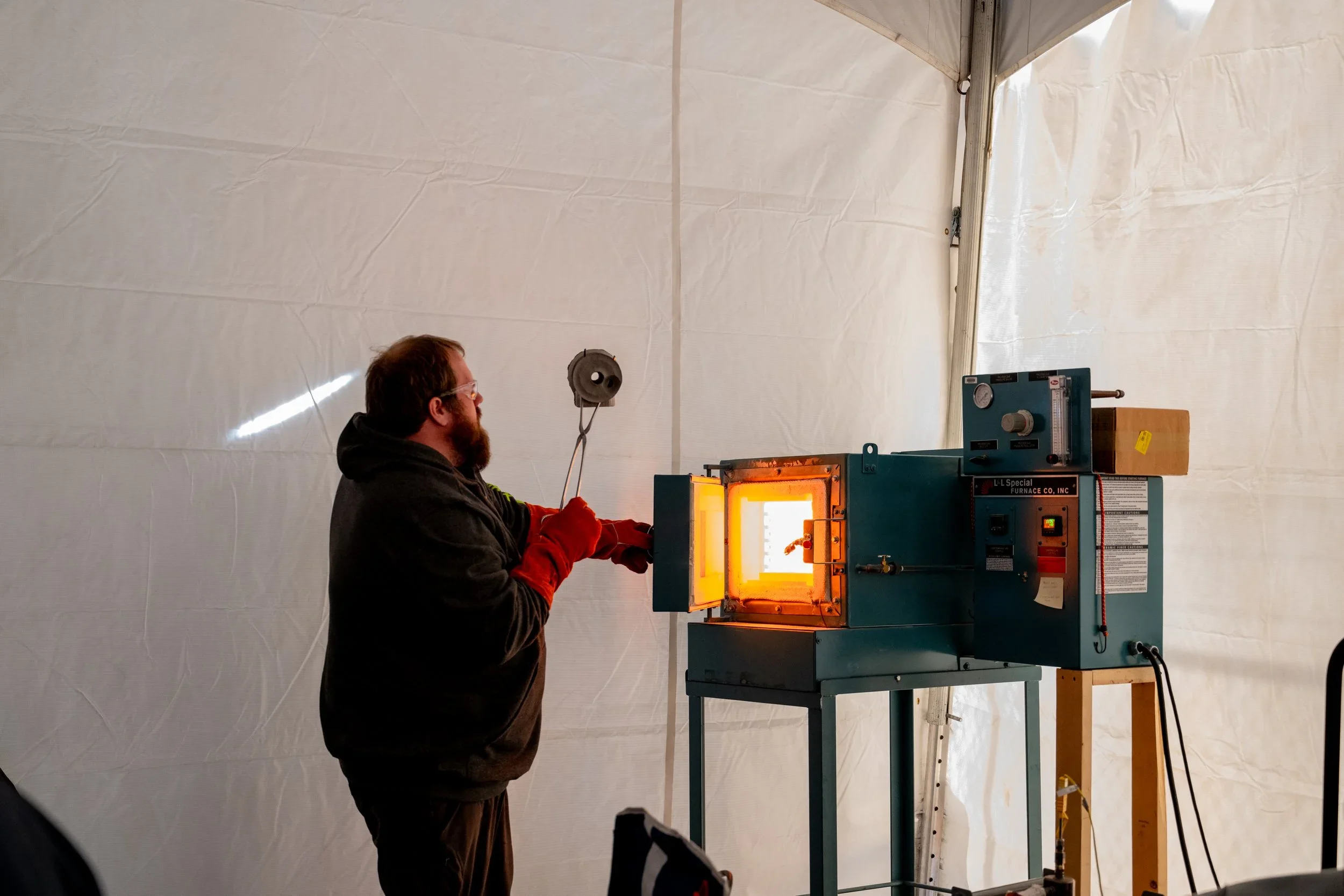 A man wearing safety goggles and red gloves standing in front of a furnace with an orange glow, working inside a large enclosed space with white fabric walls.