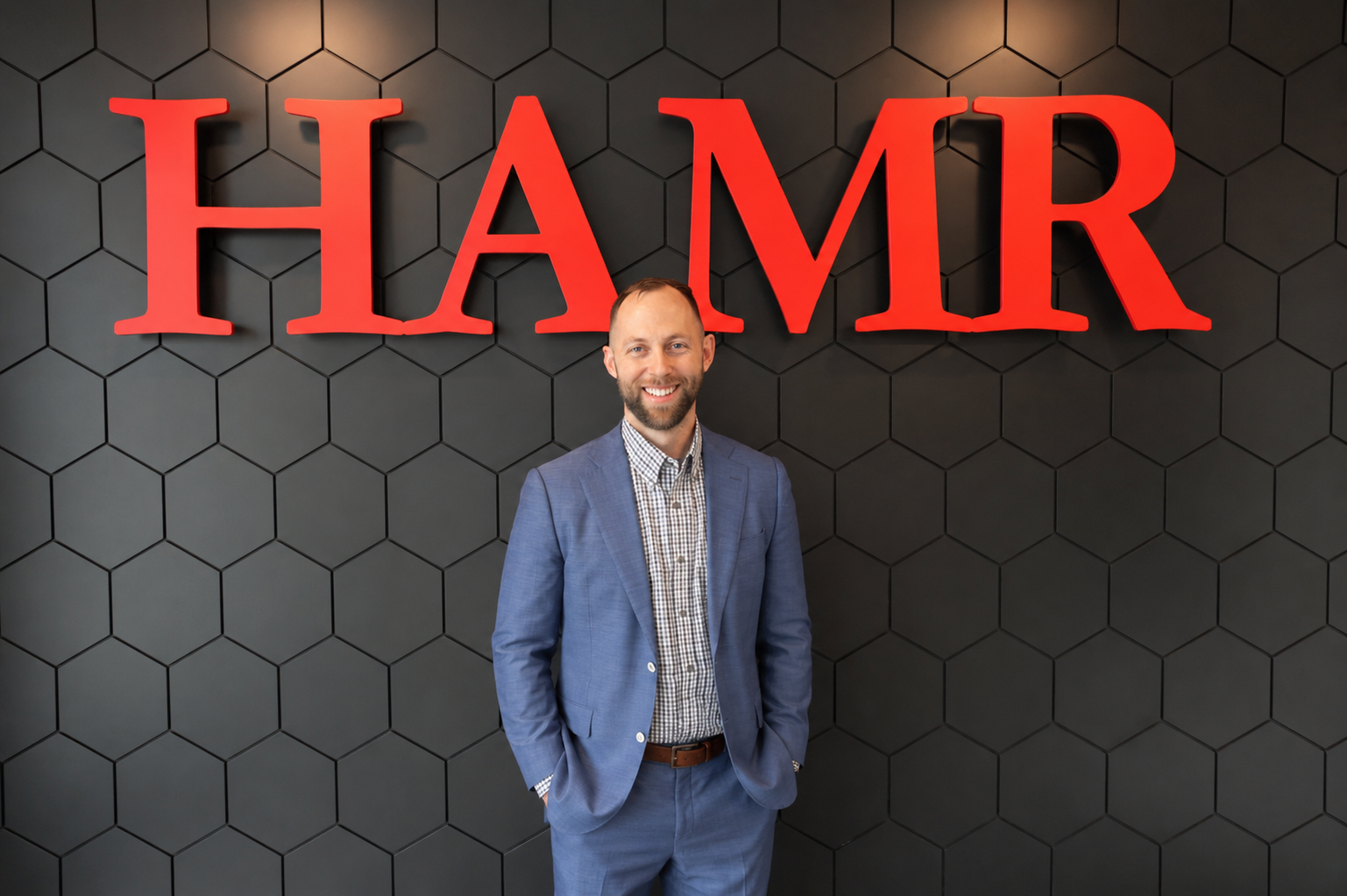 A man in a blue suit standing in front of a black wall with large red letters spelling 'HAMR'.