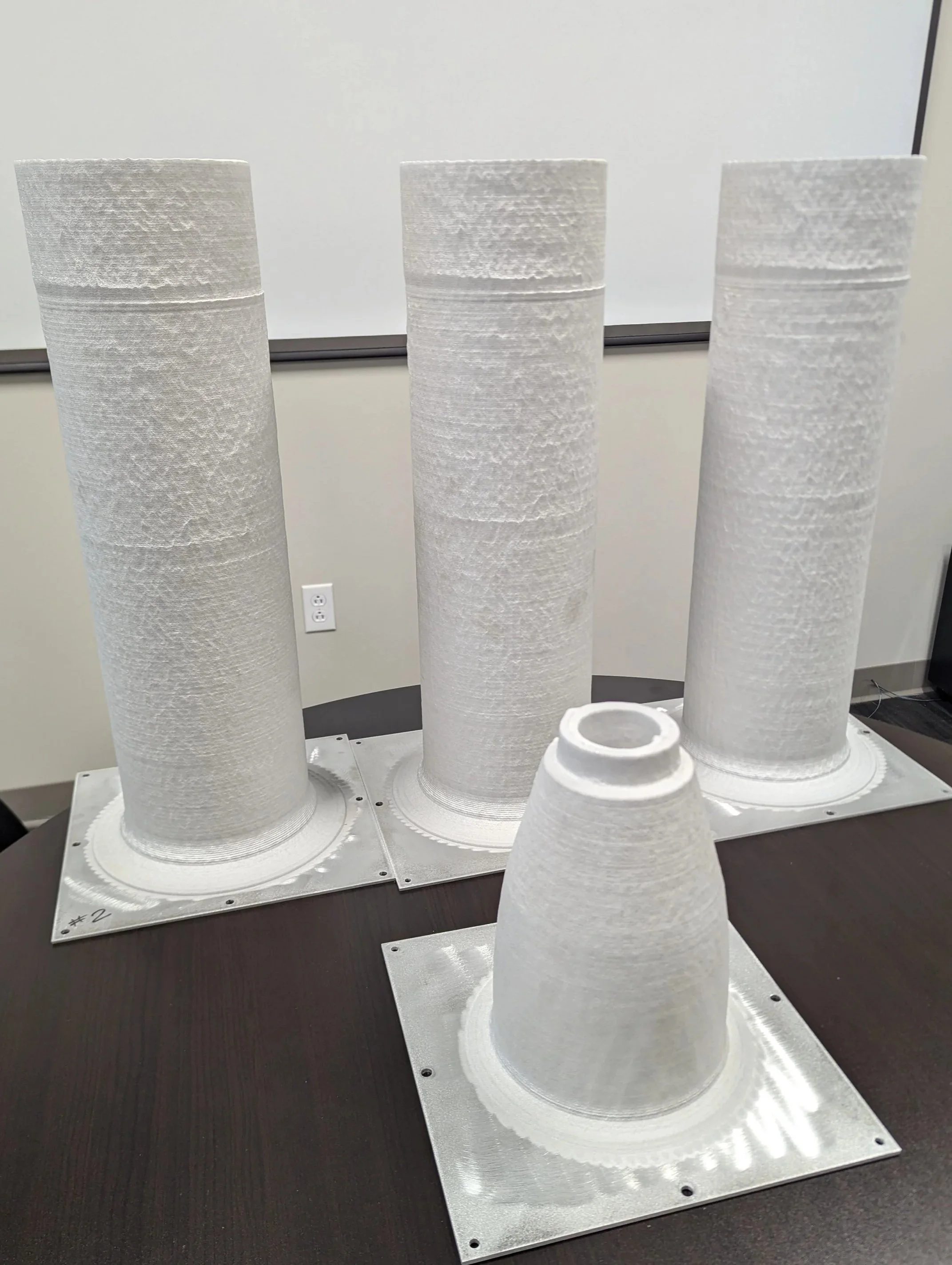 Three white 3D-printed prototype cones on metal plates, with two smaller cones in the foreground and three taller cones in the background, on a black table against a plain wall.
