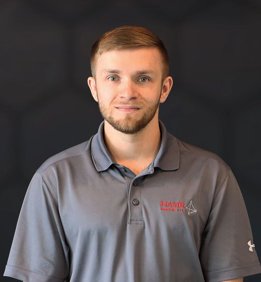 Portrait of a young man with short light brown hair and a beard, wearing a gray polo shirt with a logo on the chest, against a dark background.