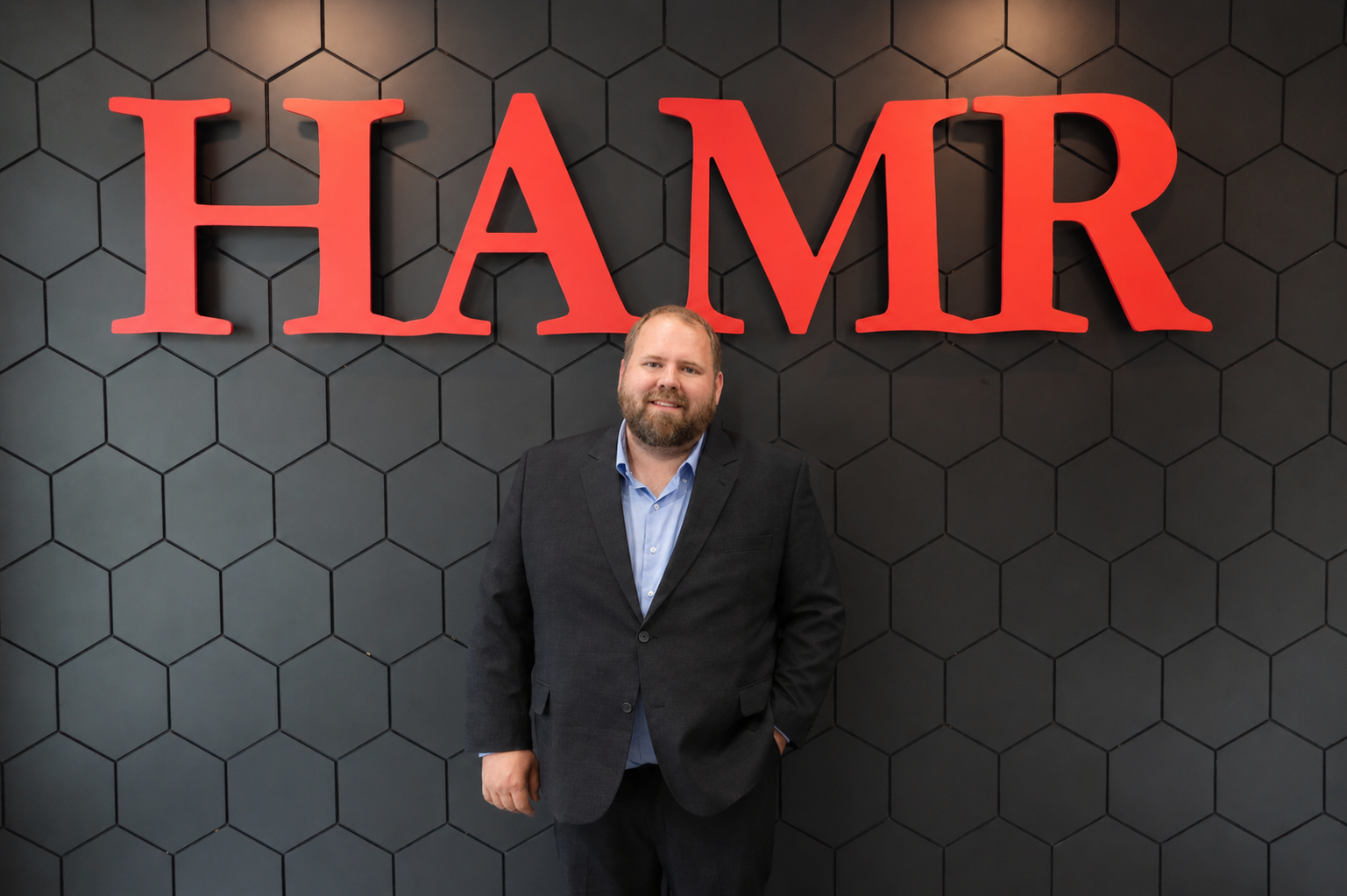 A man in a suit standing in front of a wall with a large red 'HAMR' sign, black hexagonal tiles