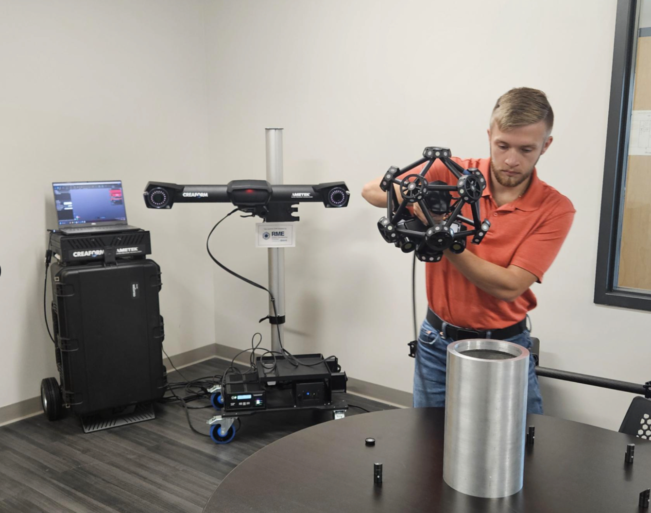 A man in an orange polo shirt is assembling a spherical structure with black segments in a room. There is a large cylinder on the table in front of him. In the background, there is a computer, a laser scanner on a stand, and electronic equipment connected with wires.