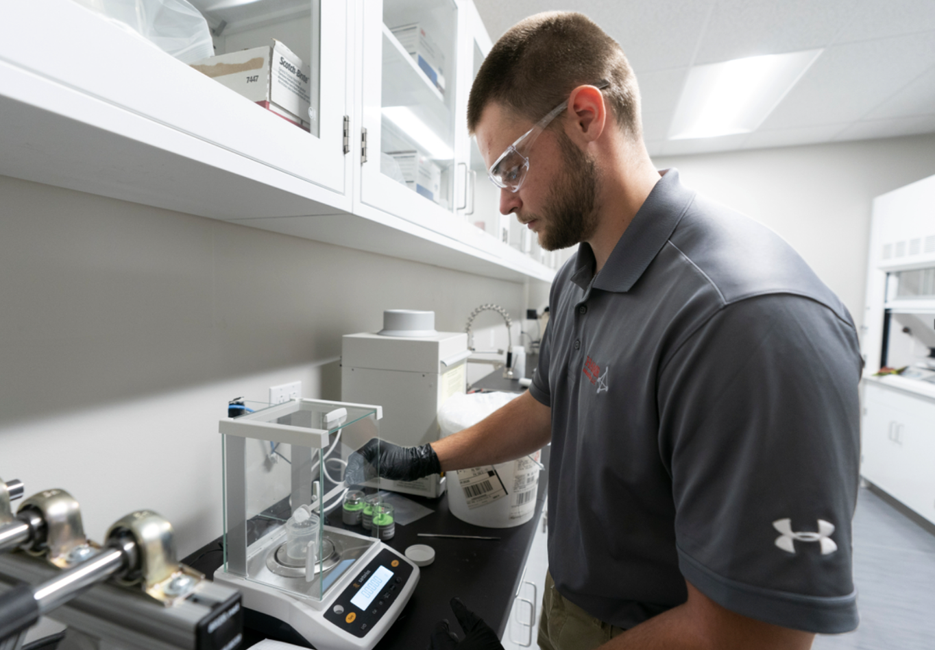 A man working in a laboratory, using a scale to measure a substance, surrounded by lab equipment and supplies.