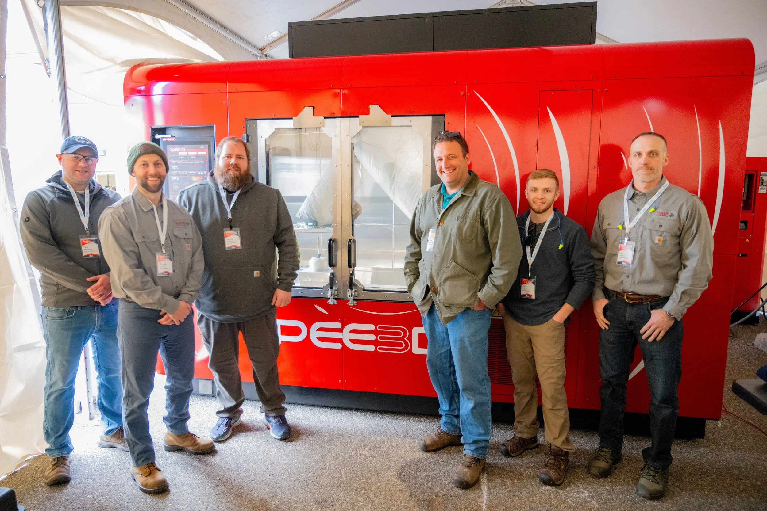 Six men standing in front of a large red 3D printing machine at an exhibition or conference.