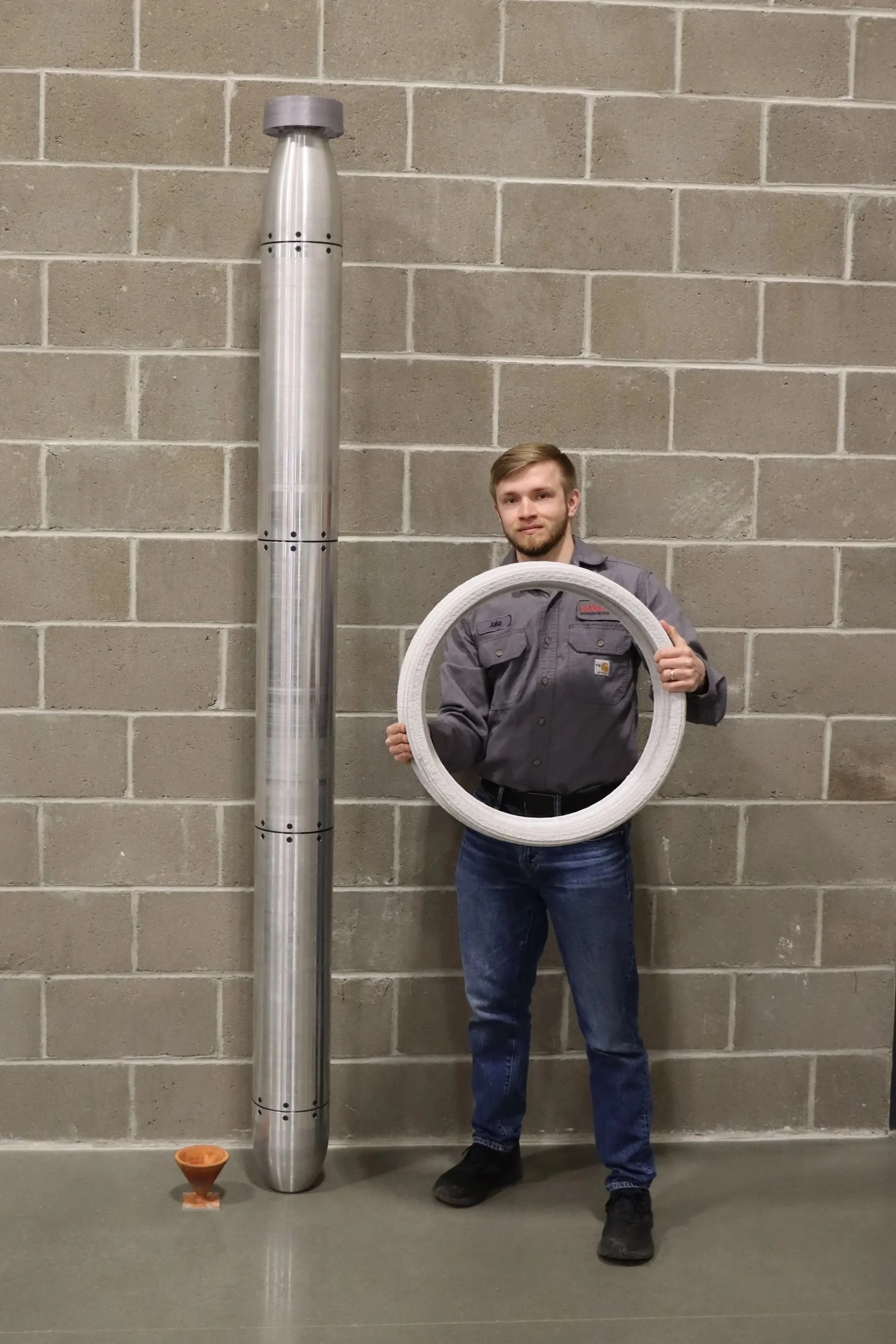 A man standing in front of a gray cinder block wall, holding a white circular ring in front of him, with a large metallic pipe to his left and a small cone shaped object on the floor nearby.