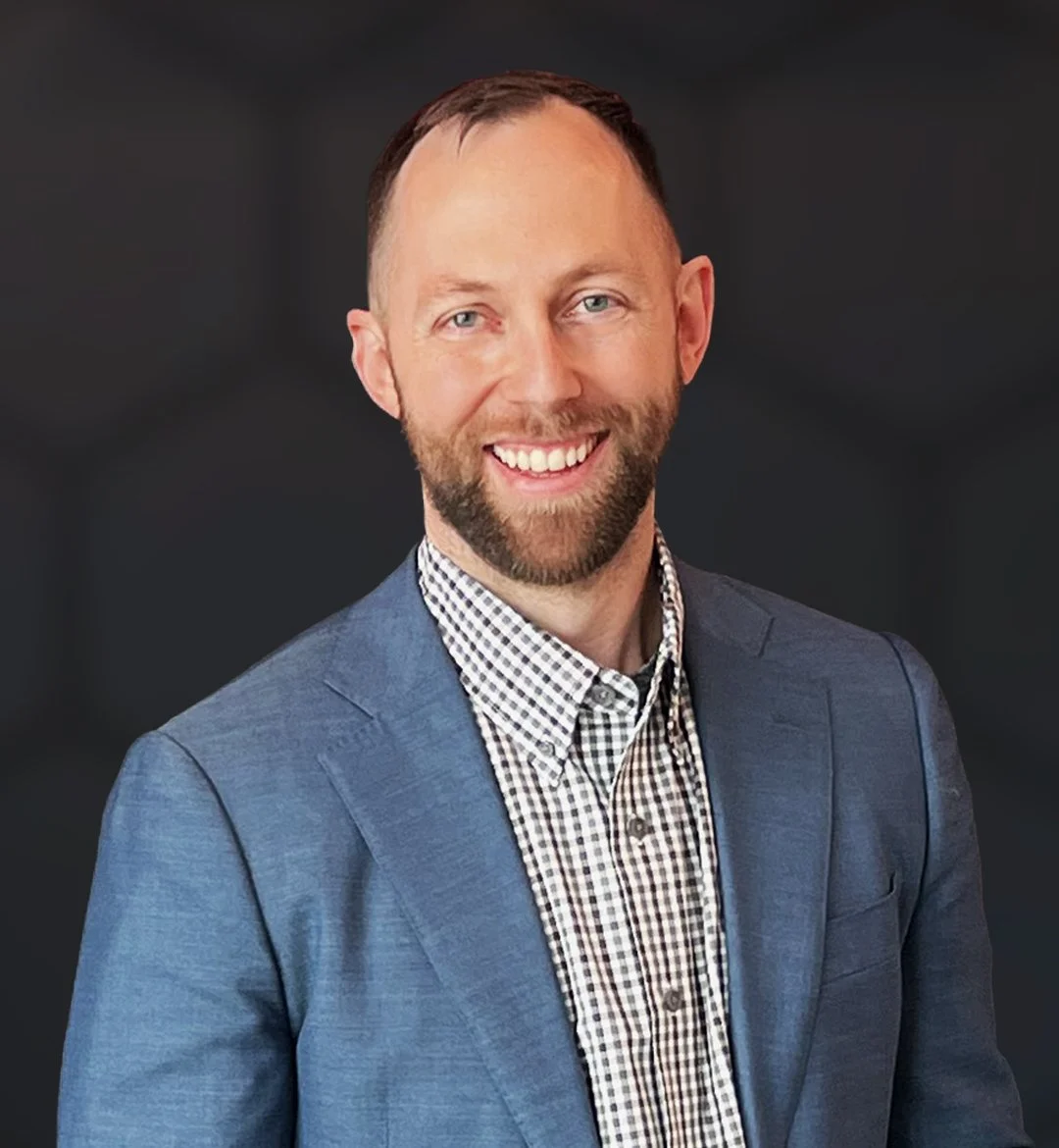 A man with light hair and beard smiling, wearing a blue blazer over a checkered shirt, against a dark background.