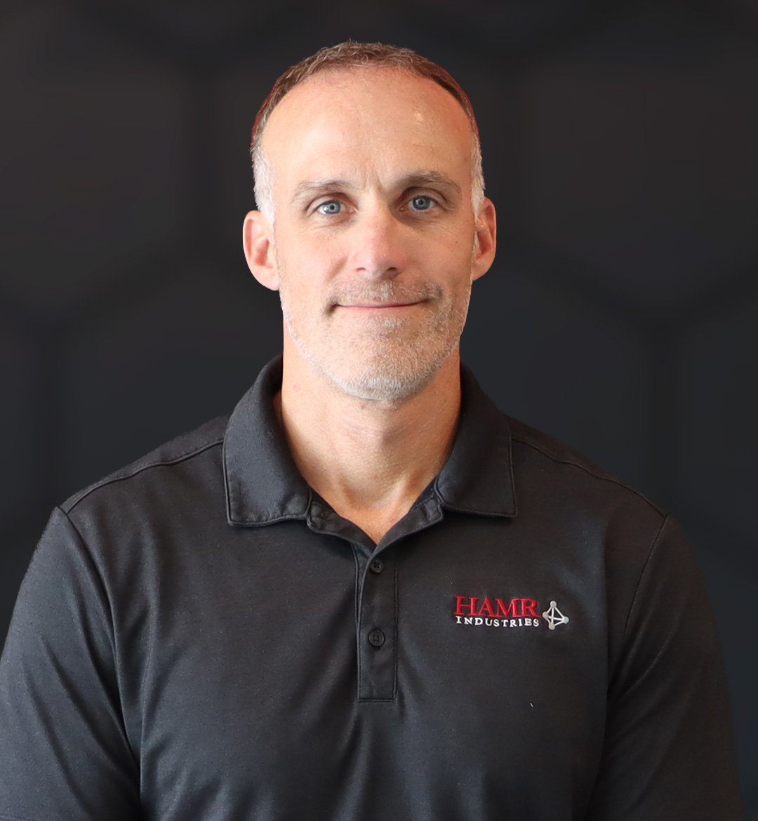 A middle-aged man with short hair and a slight beard, wearing a black polo shirt with red and white logo reading 'HAMR INDUSTRIES', posing against a dark background.