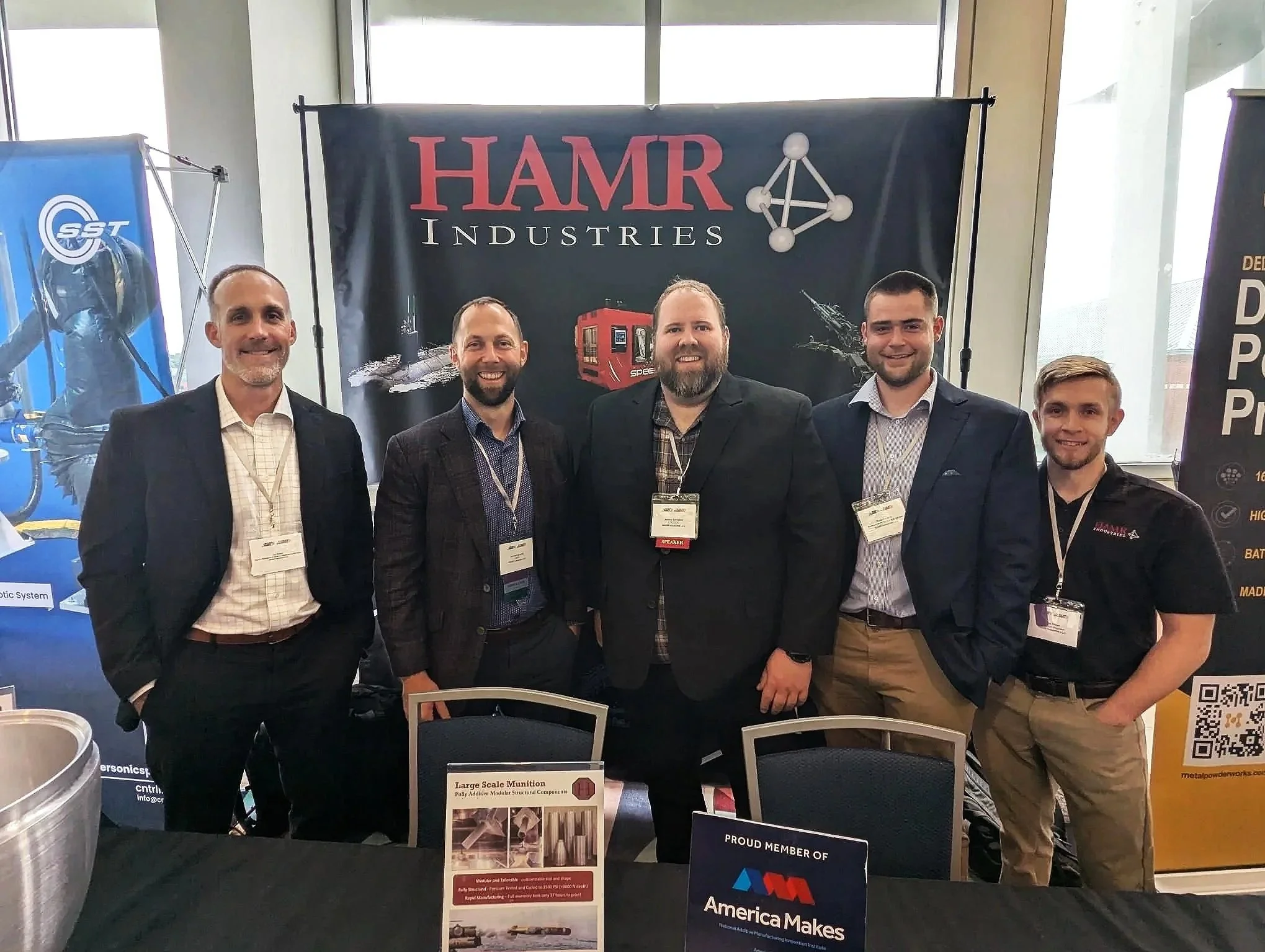 Group of five men standing together at a trade show or exhibition, smiling at the camera. Behind them is a large banner displaying the text "HAMR Industries" and a graphic of a train. There are also informational posters and signs on the table in front of them.