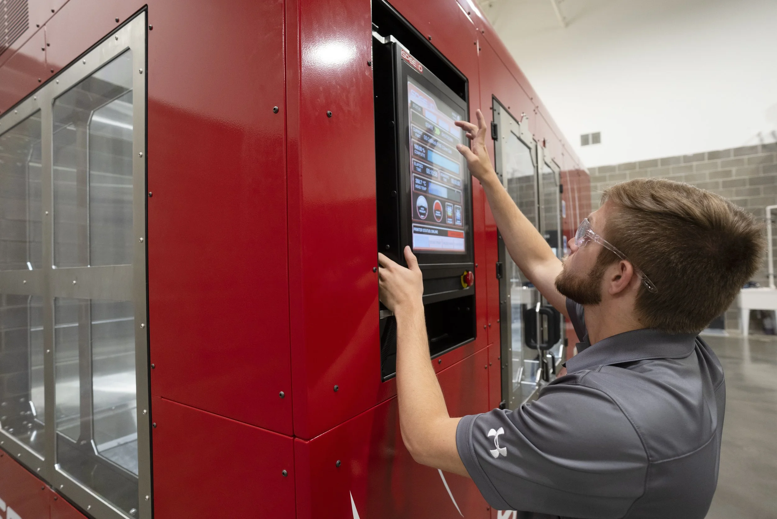 A man with safety glasses interacts with a touchscreen display on a large red machine in an industrial setting.