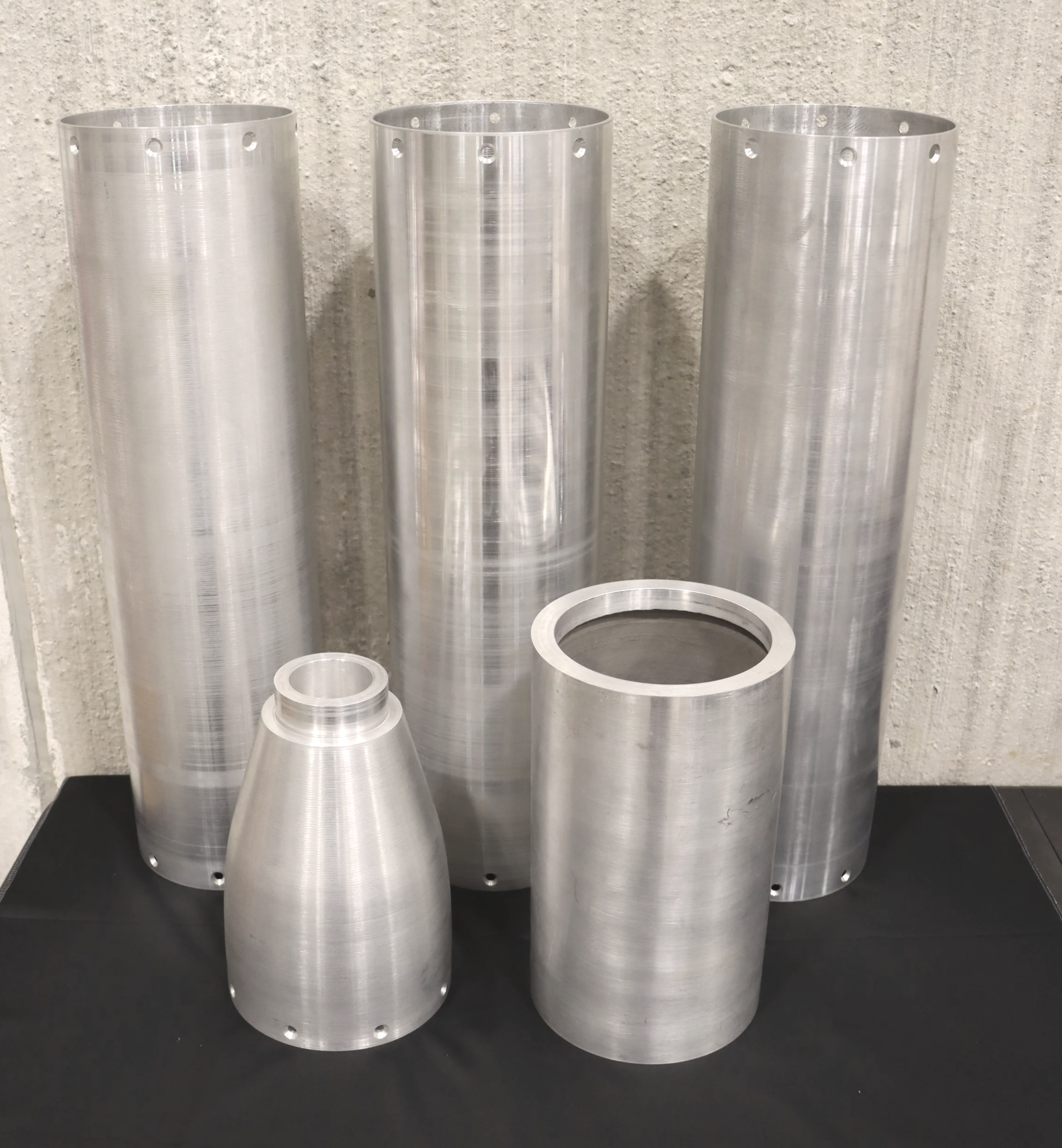 Three metallic cylinders with holes around their upper edges, placed against a textured wall, on a black surface.
