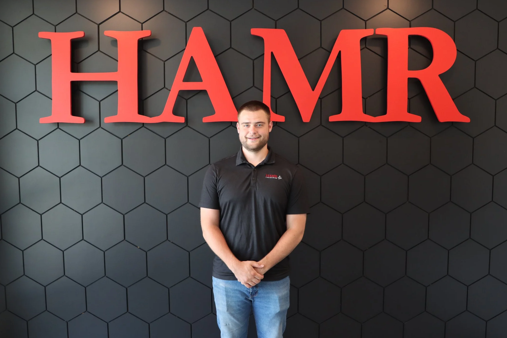 A man standing in front of a black wall with a honeycomb pattern, logo with red letters spelling 'HAMR' above his head, wearing a black polo shirt with a small logo.