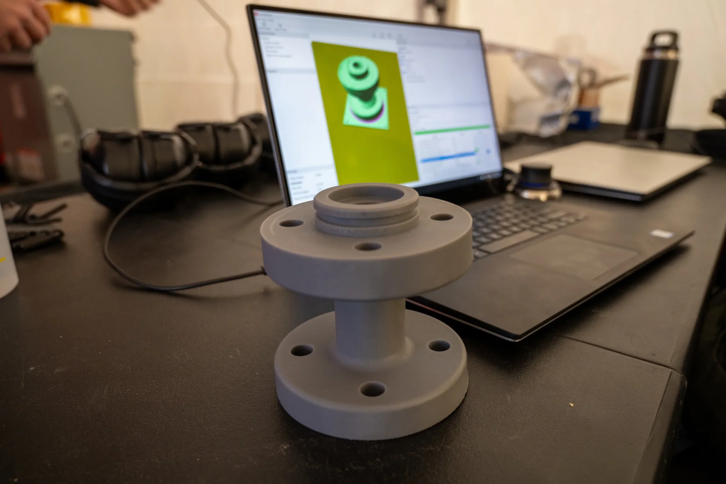 A 3D-printed object with multiple circular layers and holes, placed on a table in front of a laptop showing a 3D model on the screen, in a workspace or laboratory environment.