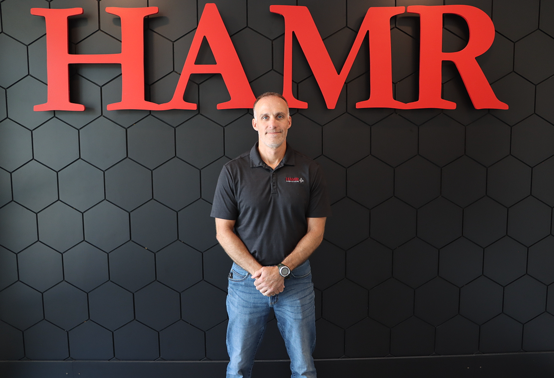 A man standing in front of a black hexagonal patterned wall with large red 3D letters spelling 'HAMR'. The man is wearing a black polo shirt with a small logo and text on the left chest, blue jeans, and a watch on his left wrist. He has short hair and is looking at the camera with a slight smile.